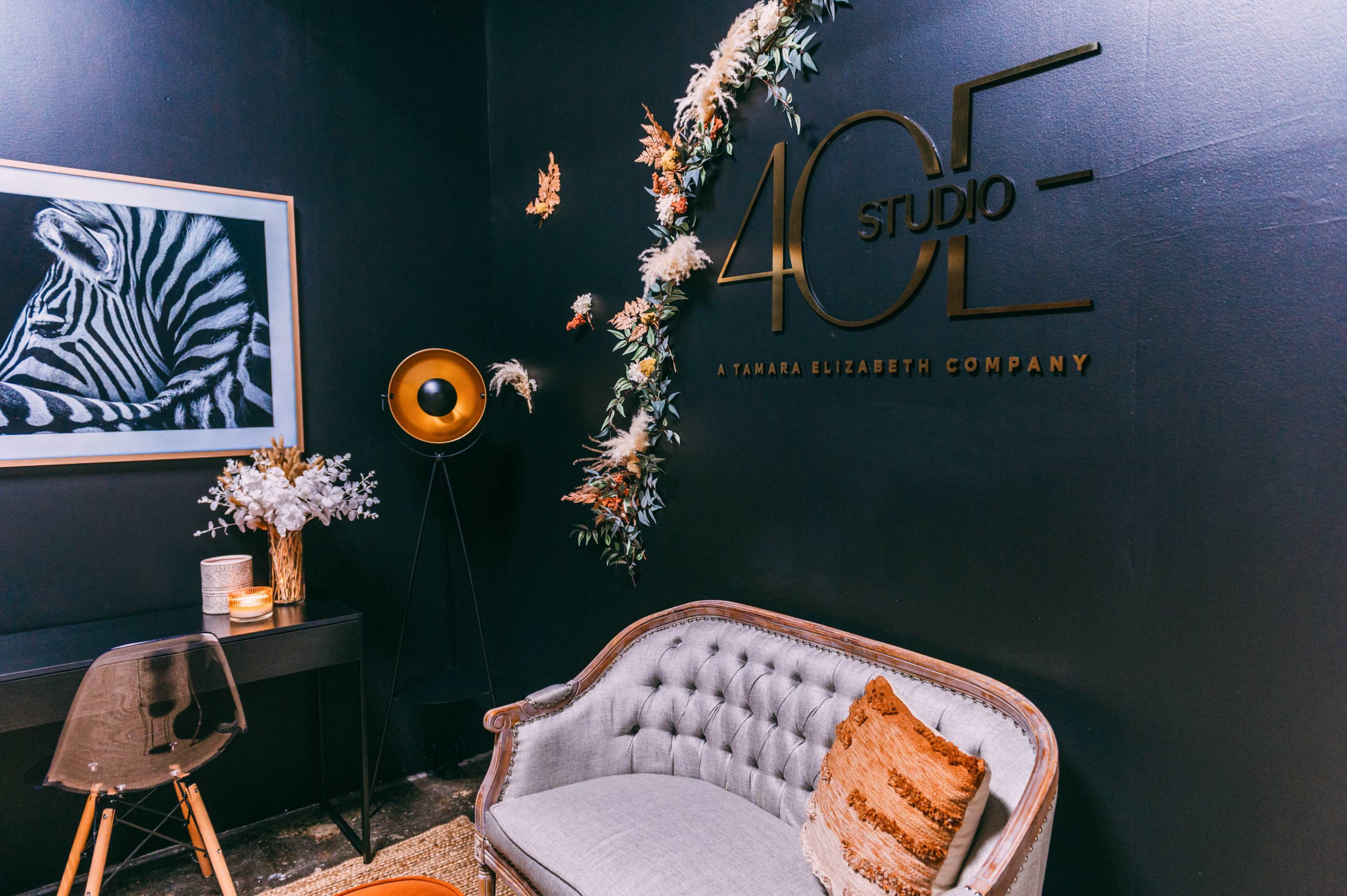 A stylish interior featuring a decorative wall with a sign that reads "40E STUDIO," a plush couch, a modern lamp, and a floral arrangement.