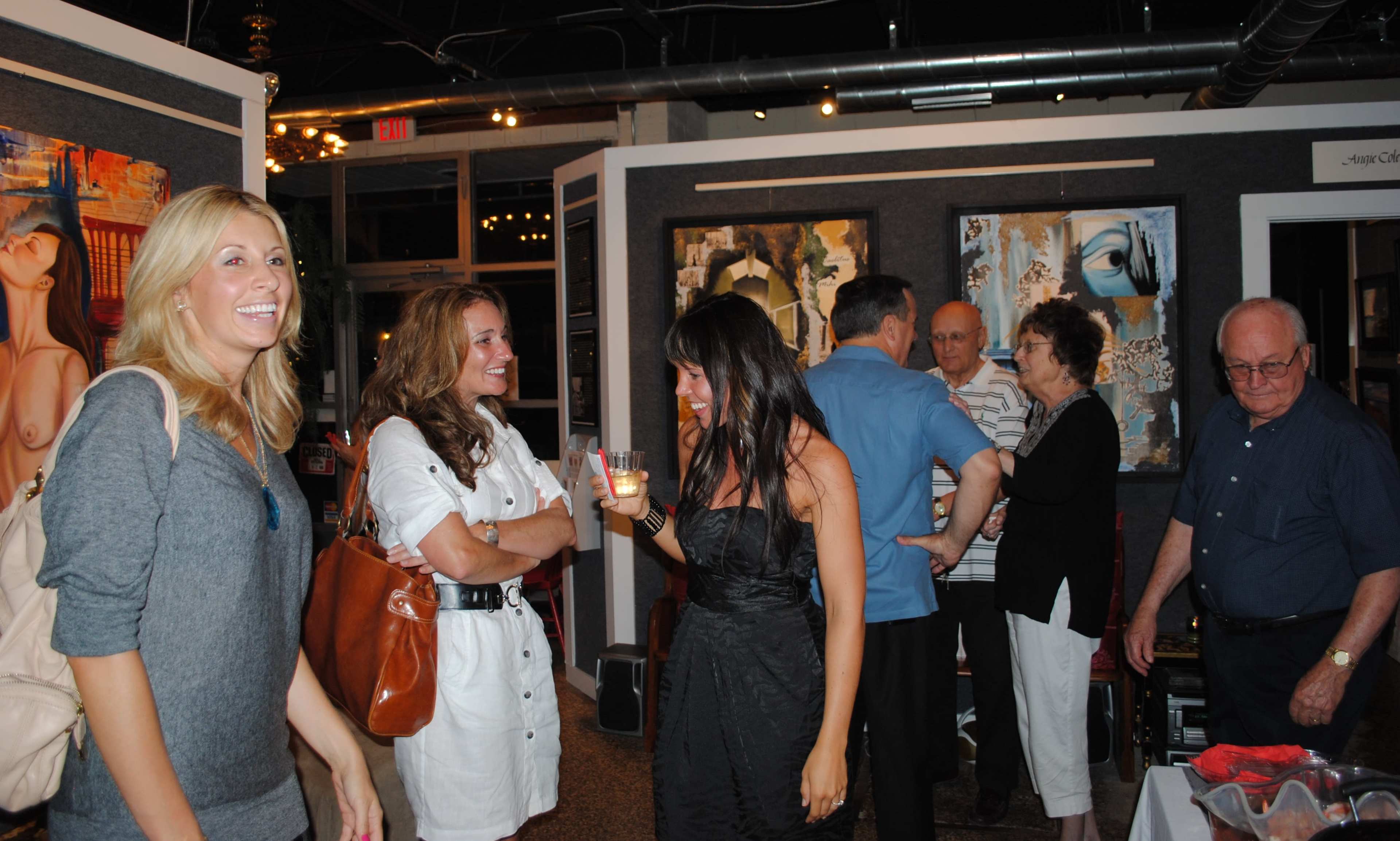 A group of people socializes at an art gallery exhibition, with some interacting and others observing the artwork.