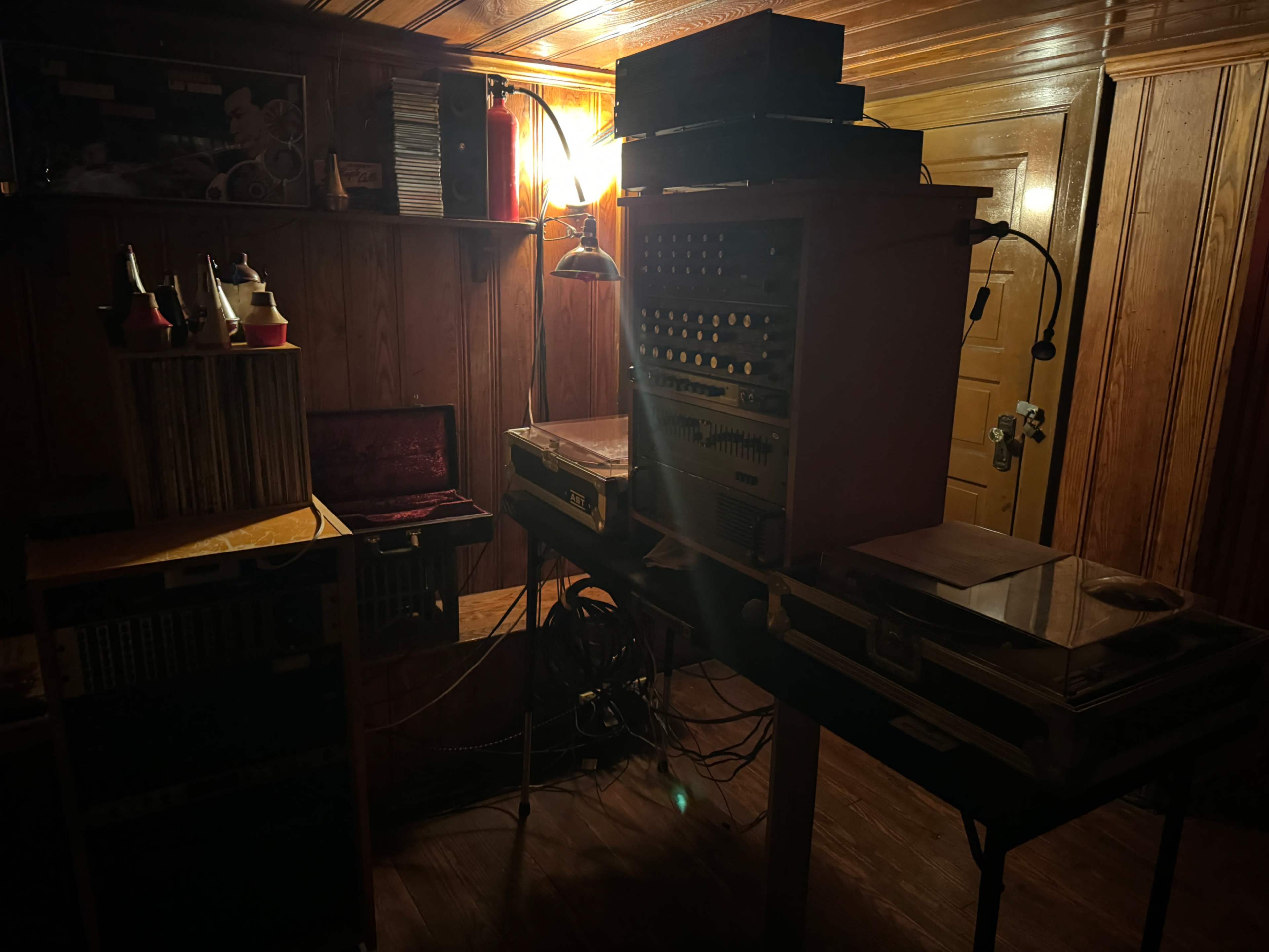A dimly lit room with wooden paneling, featuring a vintage sound system, record players, and various audio equipment arranged on tables.
