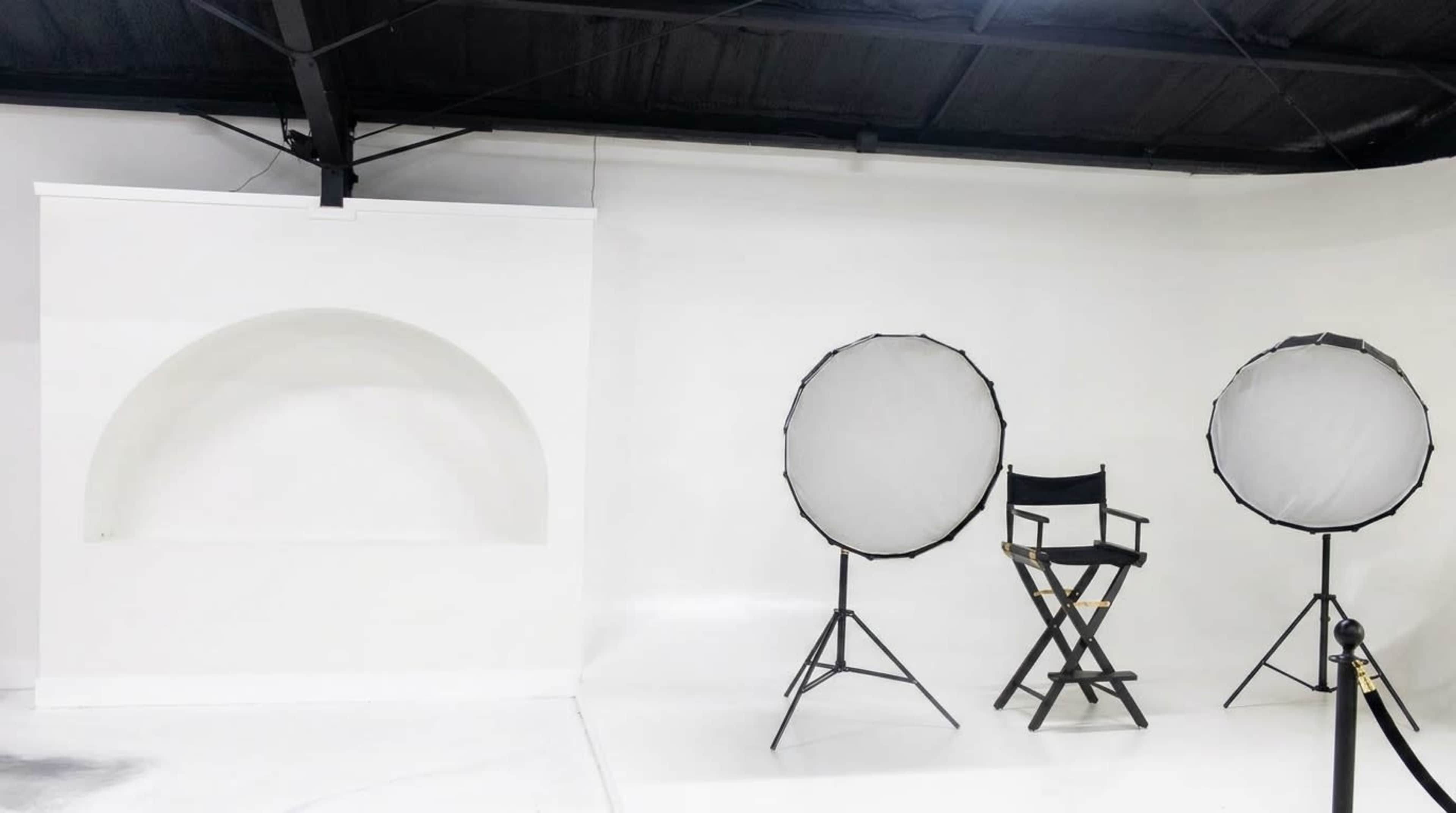 The image shows a minimalist studio space with a white backdrop, a director's chair, and two softbox lighting setups.