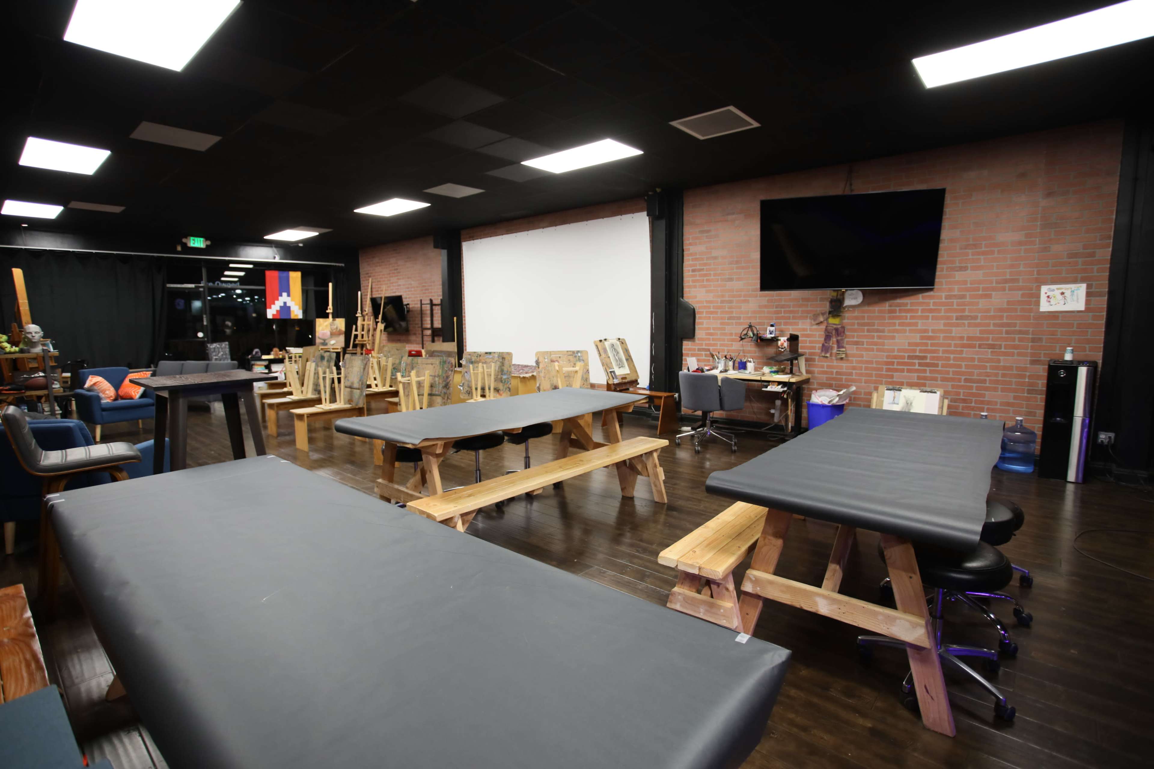 A spacious interior with several wooden tables and chairs in a studio, featuring a large blank wall, a television, and various art supplies in the background.