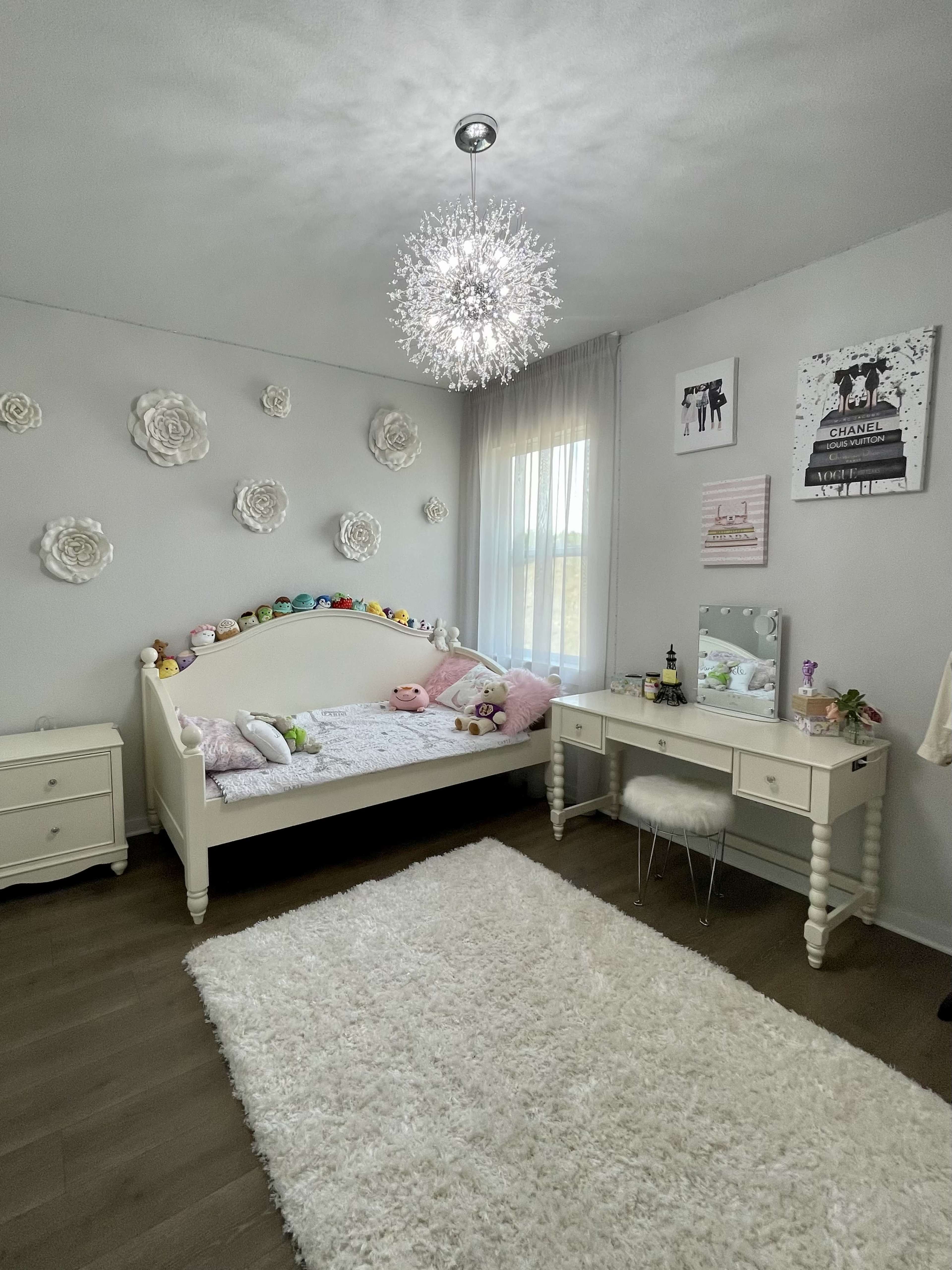 The image shows a softly lit bedroom featuring a white bed with decorative pillows, a small vanity with a mirror, and a large, fluffy white area rug on the wooden floor.