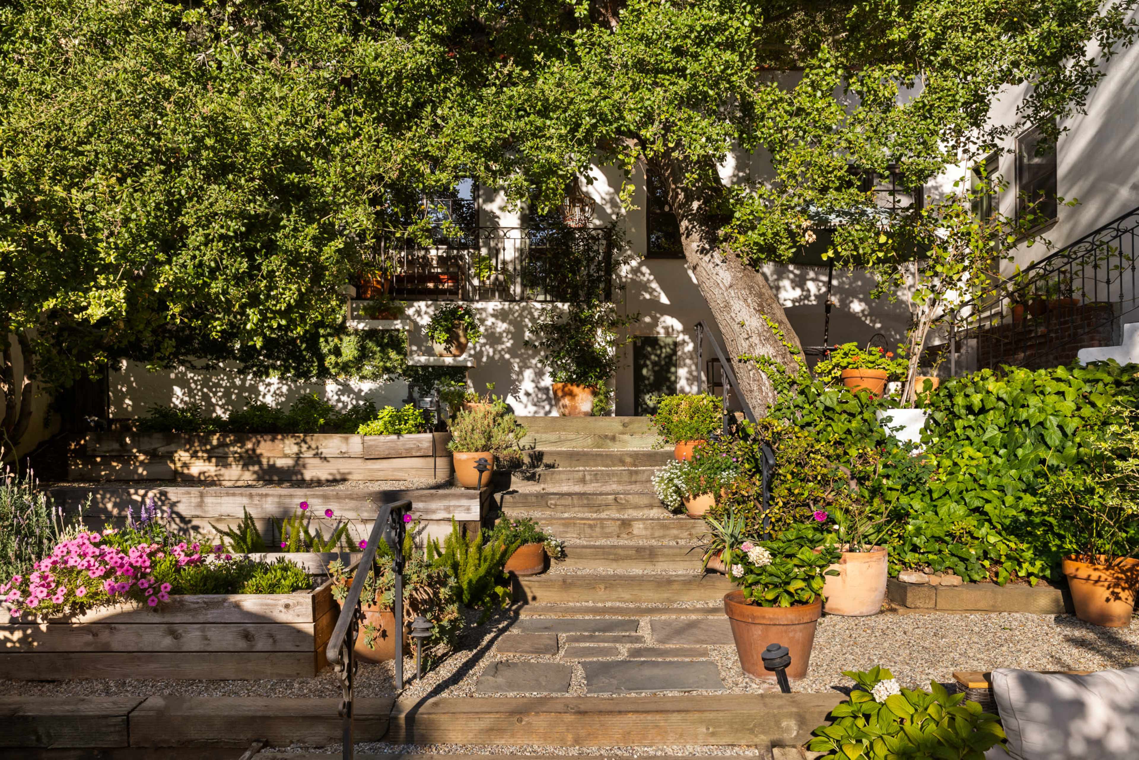 The image shows a stone staircase leading to a white building, surrounded by various potted plants and garden beds filled with colorful flowers.