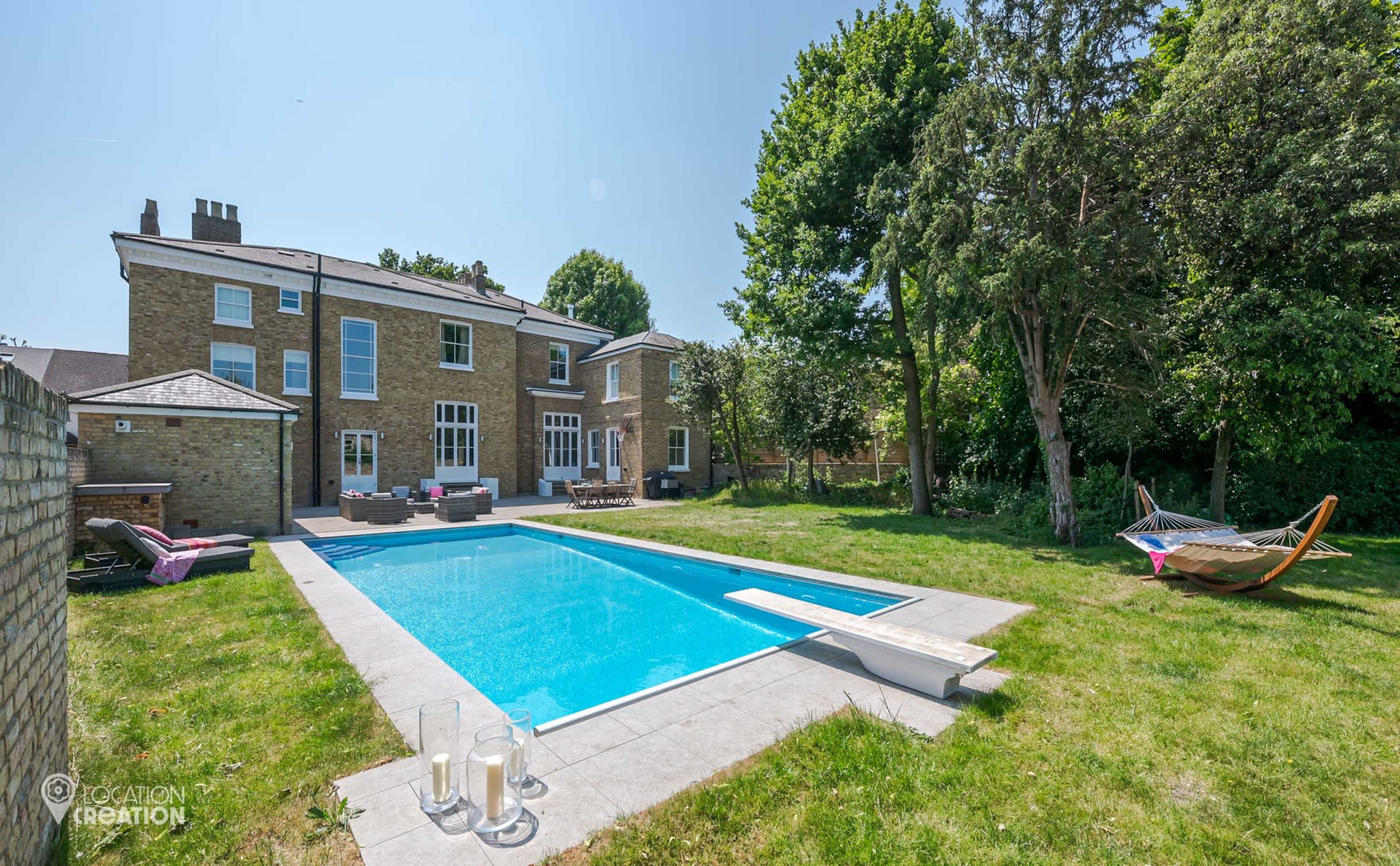 The image shows a spacious backyard featuring a swimming pool, a lounge area with outdoor furniture, and a hammock, all surrounded by greenery and a large brick house.