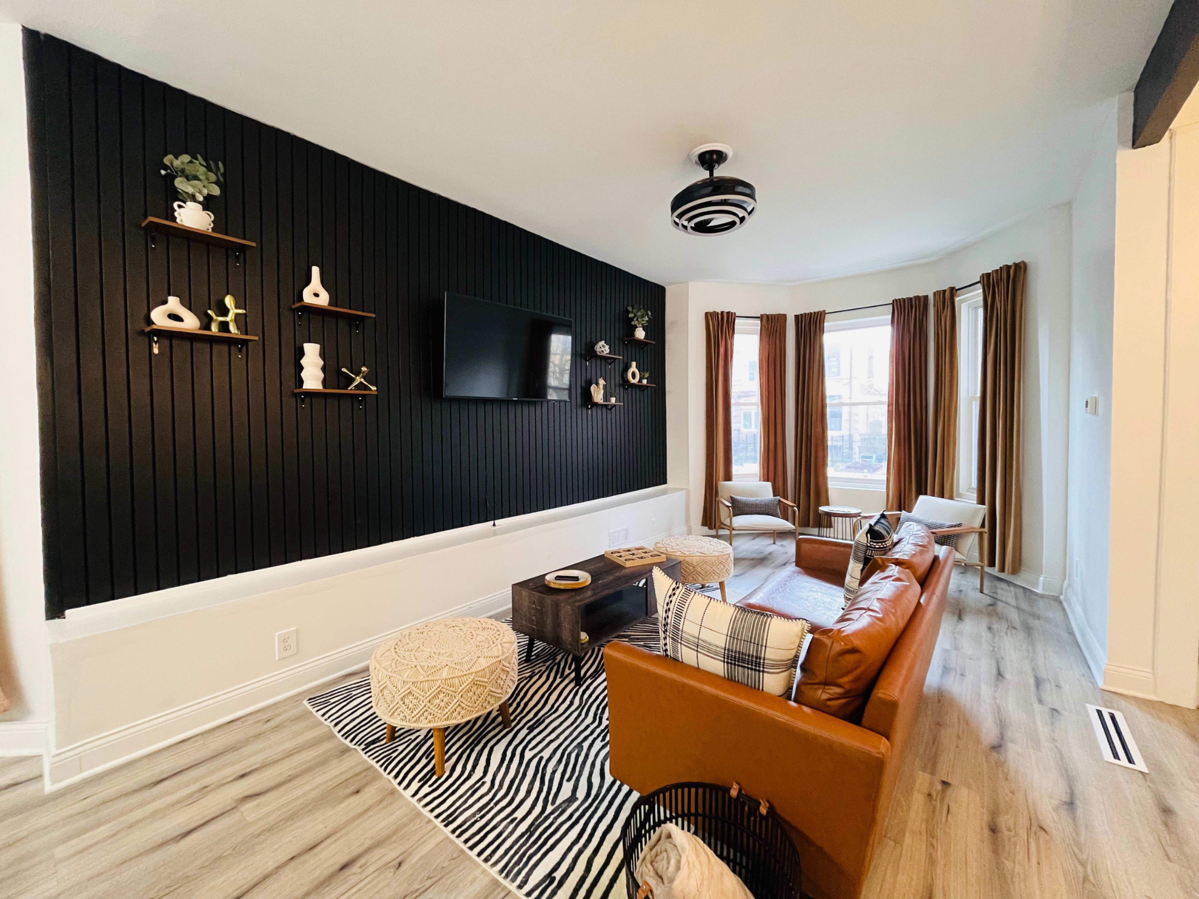The living room features a black accent wall with decorative shelves, a flat-screen TV, a brown leather sofa, and large windows with brown curtains.