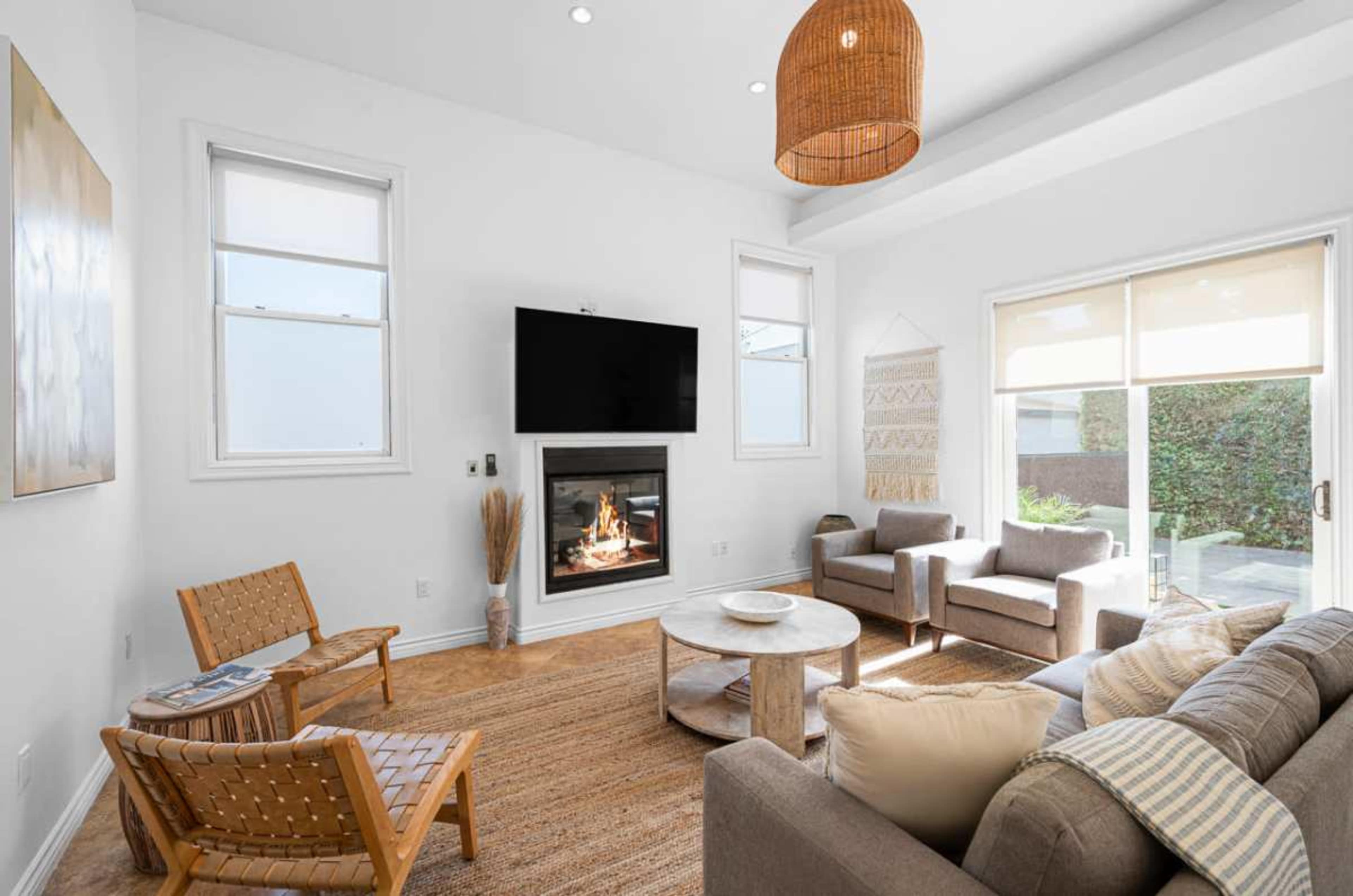 The image depicts a well-lit living room featuring a modern fireplace, a television mounted on the wall, two armchairs, and a round coffee table on a woven rug.