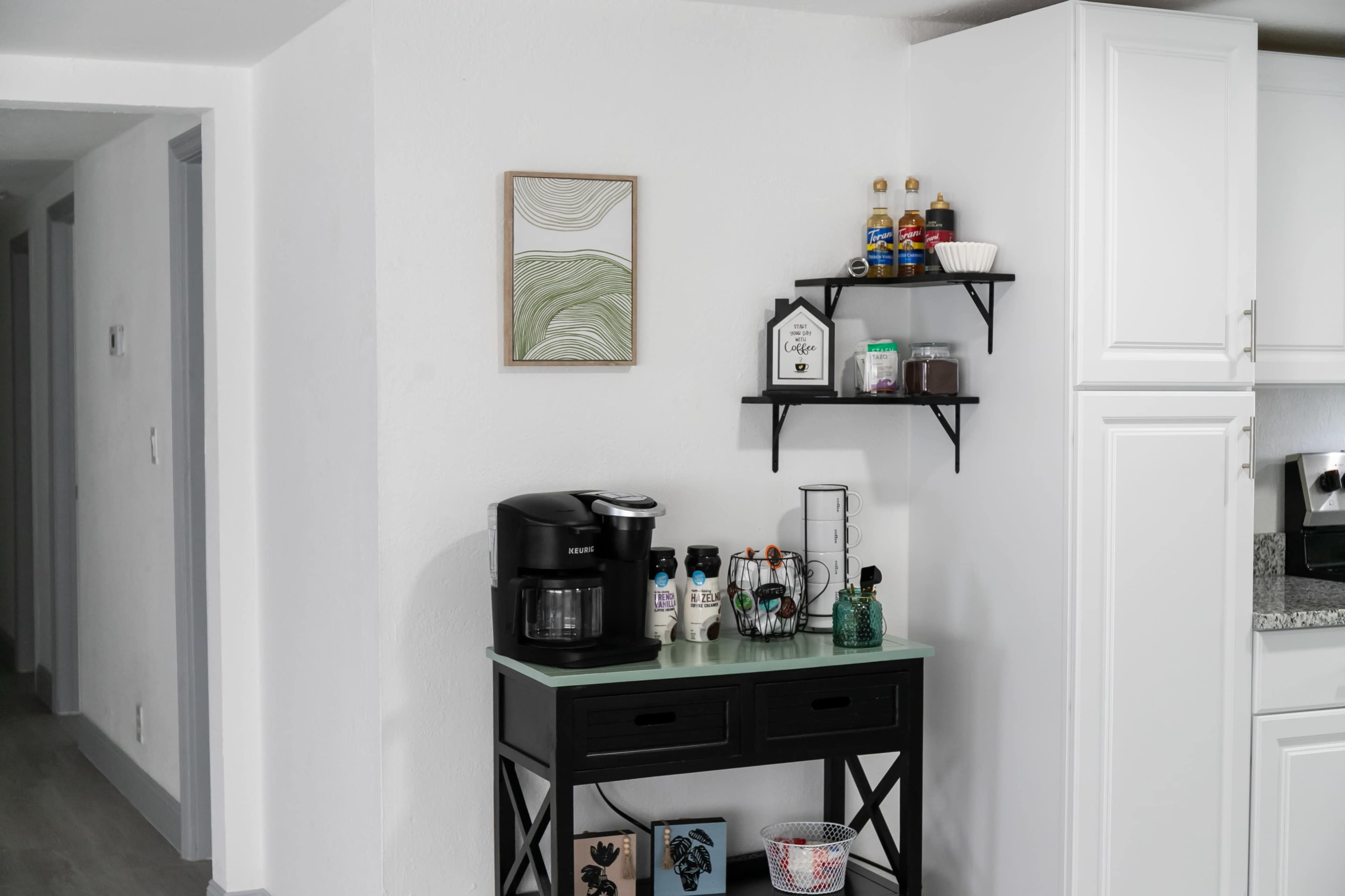 A black coffee maker is positioned on a dark console table beneath a wall-mounted shelf displaying various condiments and a small framed piece of artwork.