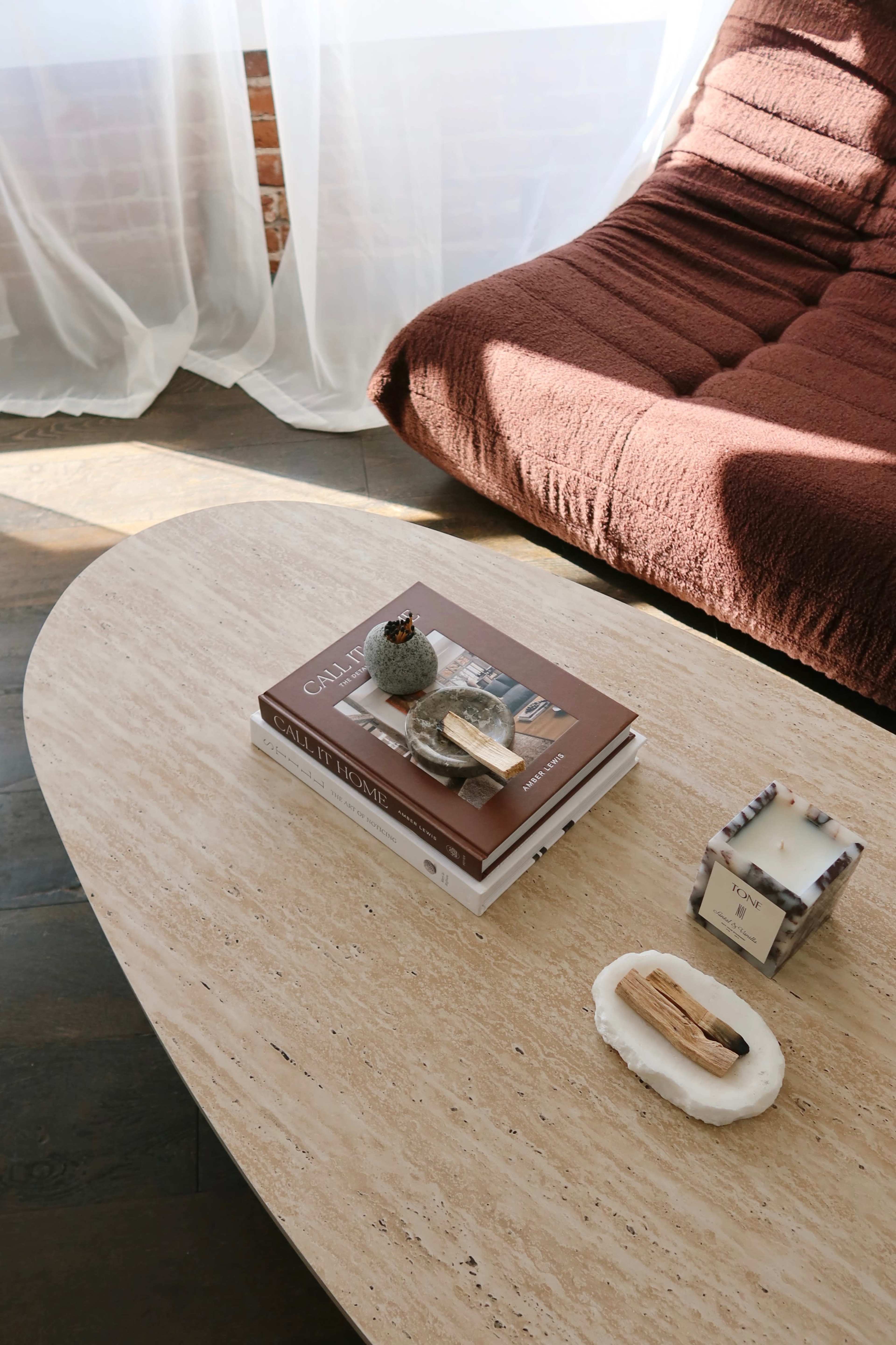 A coffee table topped with a few books, a decorative object, and a candle sits in a sunlit room with a sofa nearby.