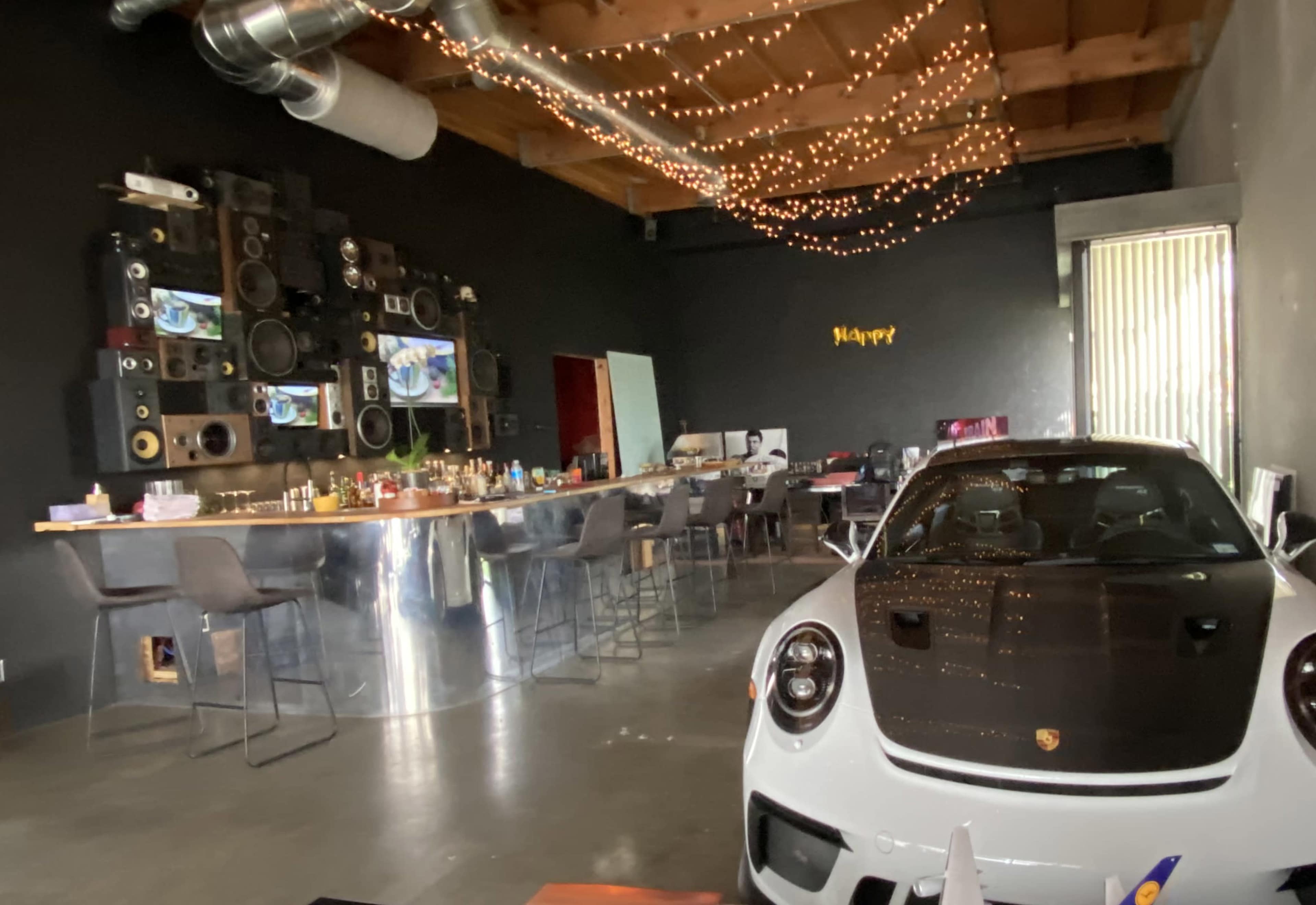 A white Porsche sports car is parked in a modern, stylish room featuring a long bar, high stools, and a wall adorned with large speakers and fairy lights.