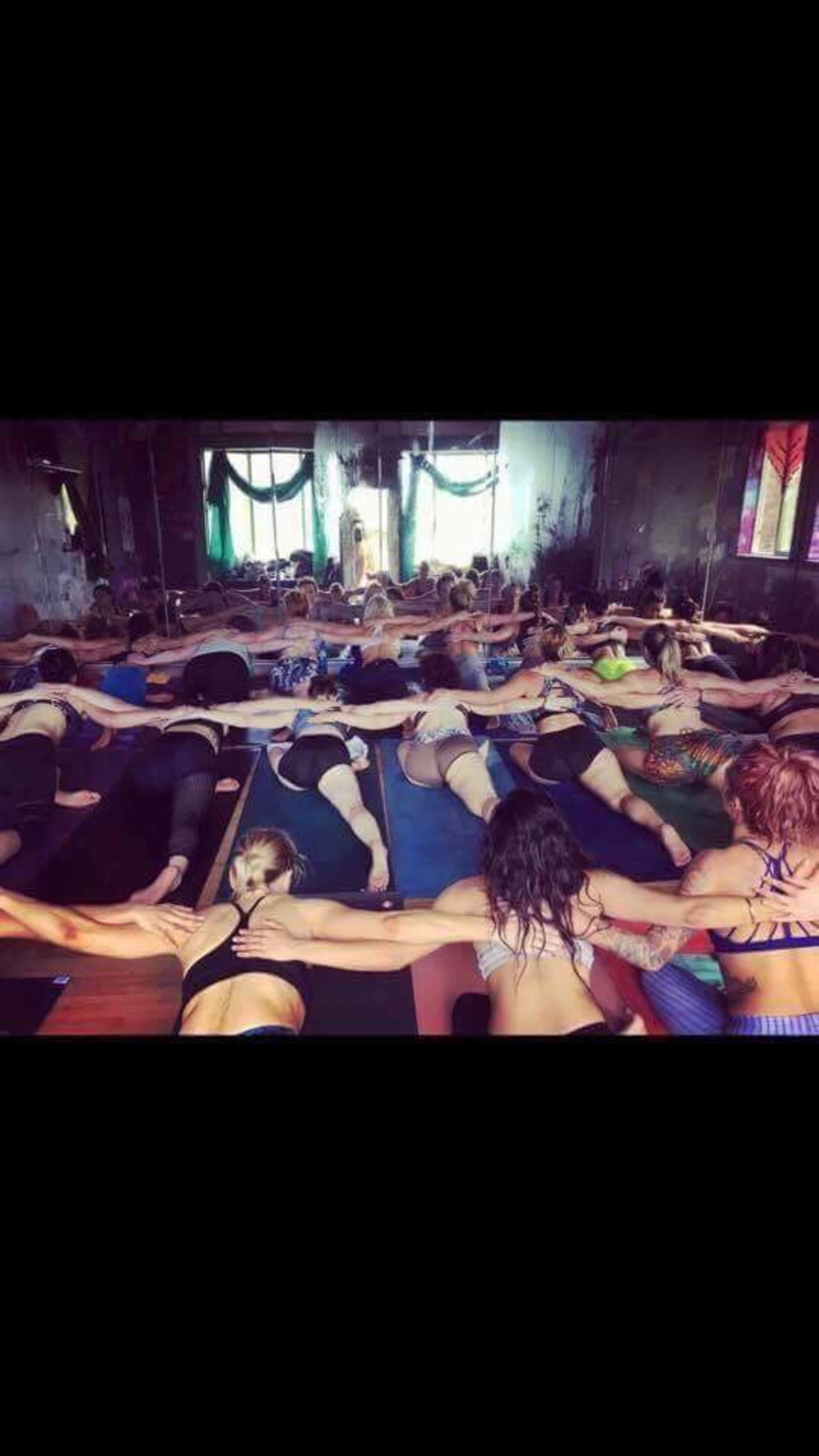A large group of people practice yoga in a dimly lit studio, connected by holding hands while lying on mats.
