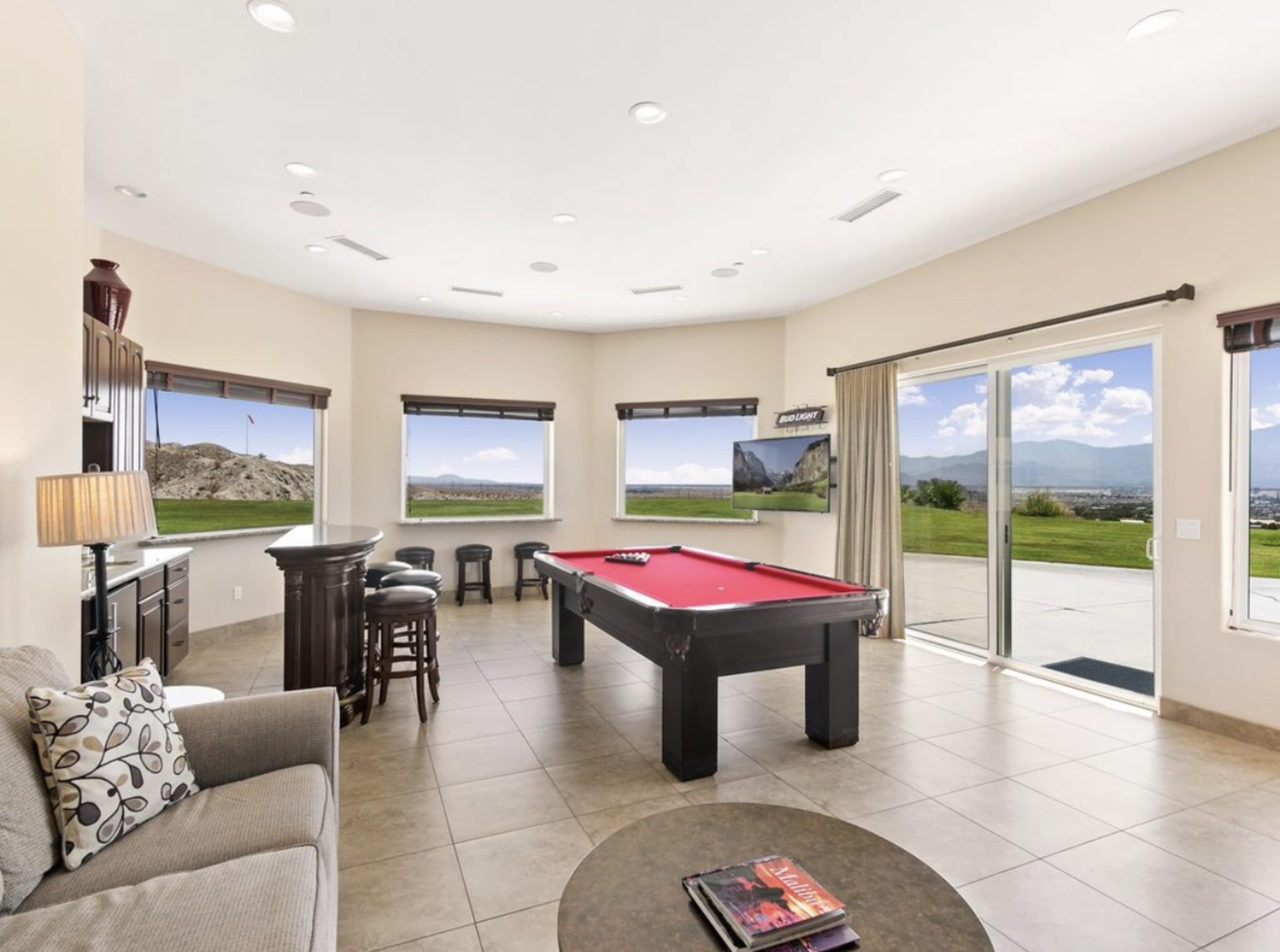 A game room with a billiards table, a seating area, and large windows offering a view of the outdoors.