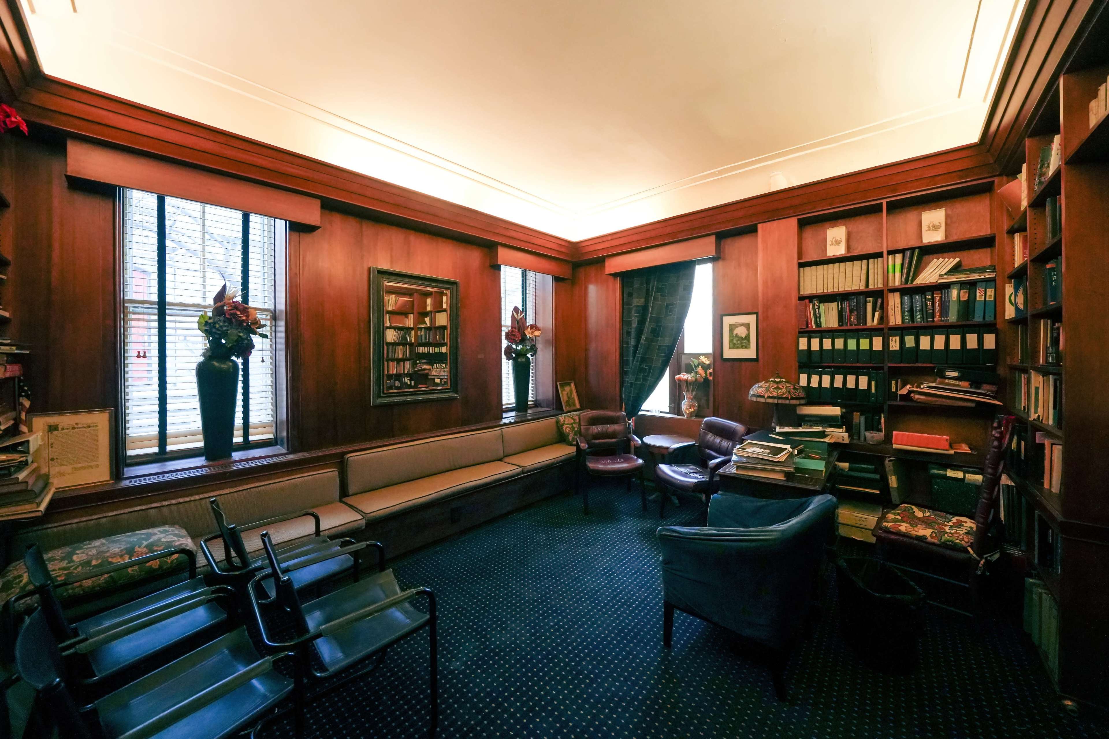 A cozy library with wooden walls, a sofa along one side, two armchairs, and bookshelves filled with books.