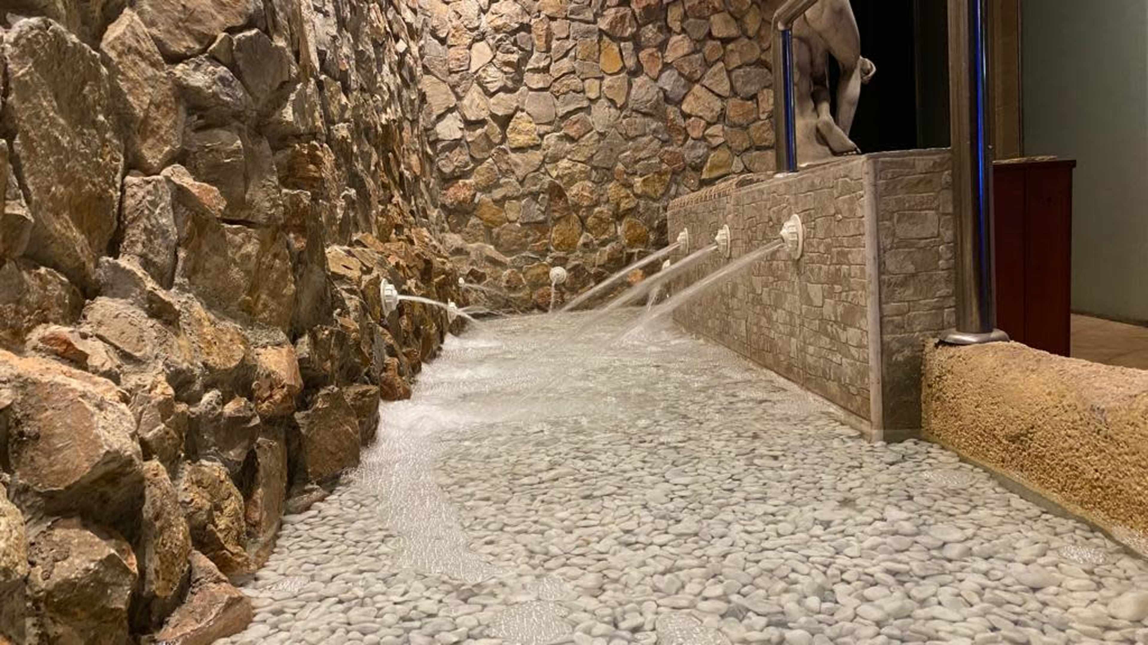 Water flows from wall-mounted jets into a shallow pebble-lined stream, surrounded by a stone wall.