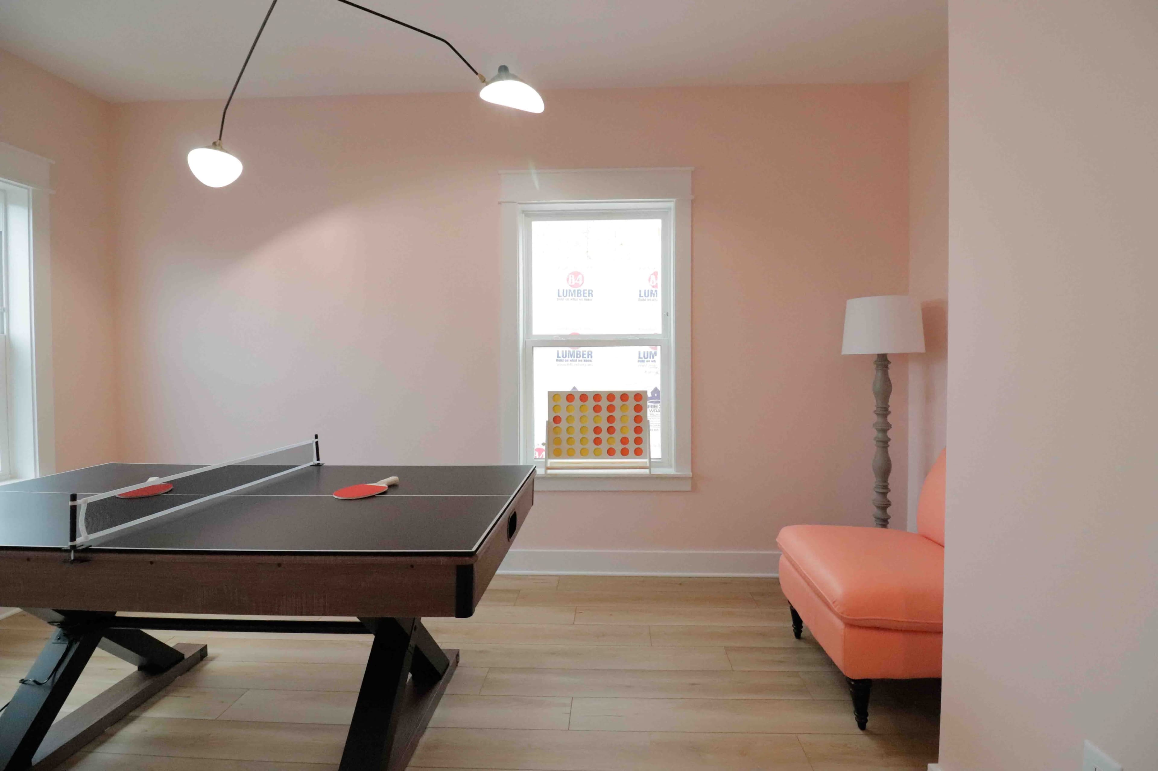 A light-colored room featuring a ping pong table with paddles and a peach-colored chair beside a window.