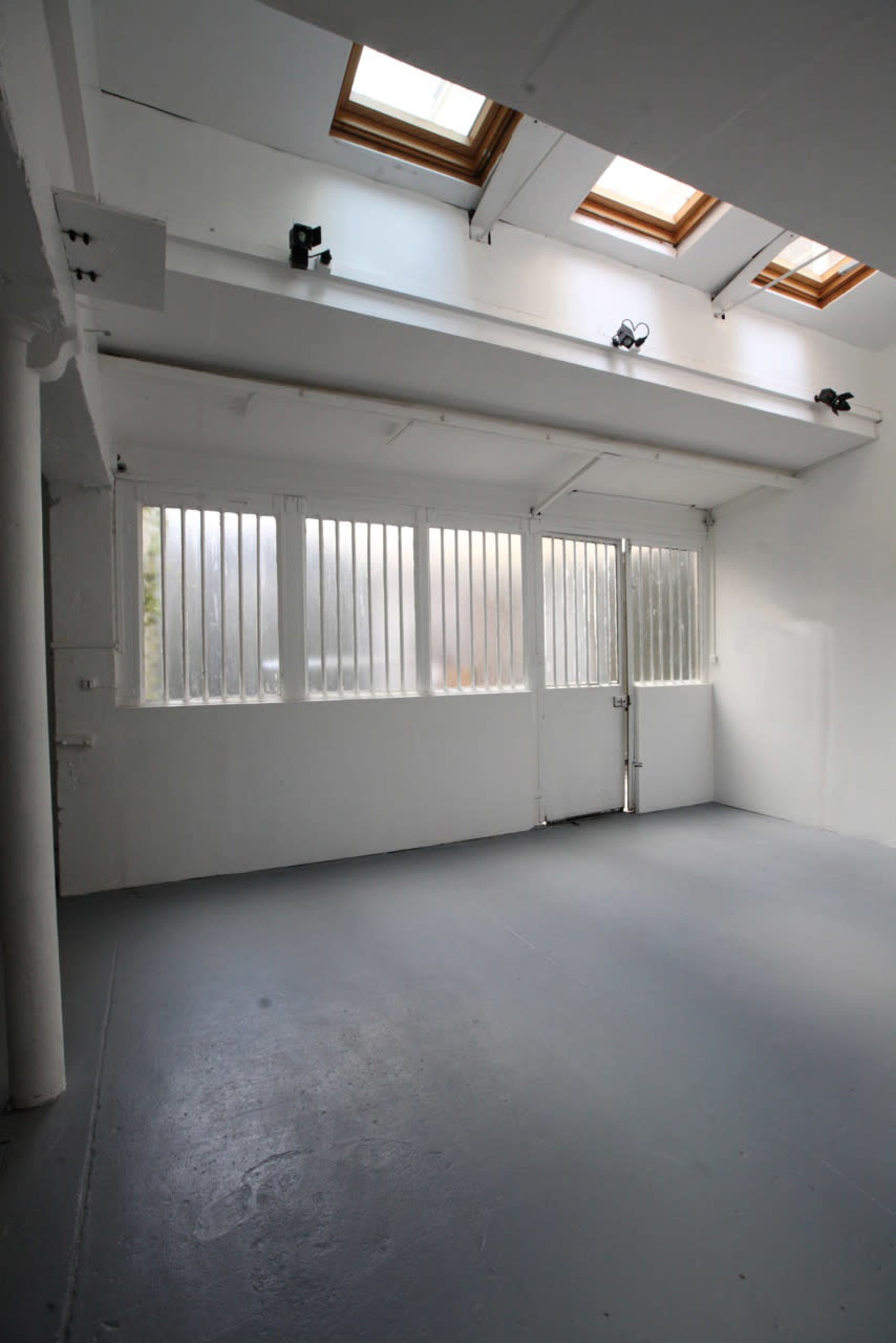 A spacious, empty room with white walls, high ceilings, and windows with vertical slats allowing light to enter.