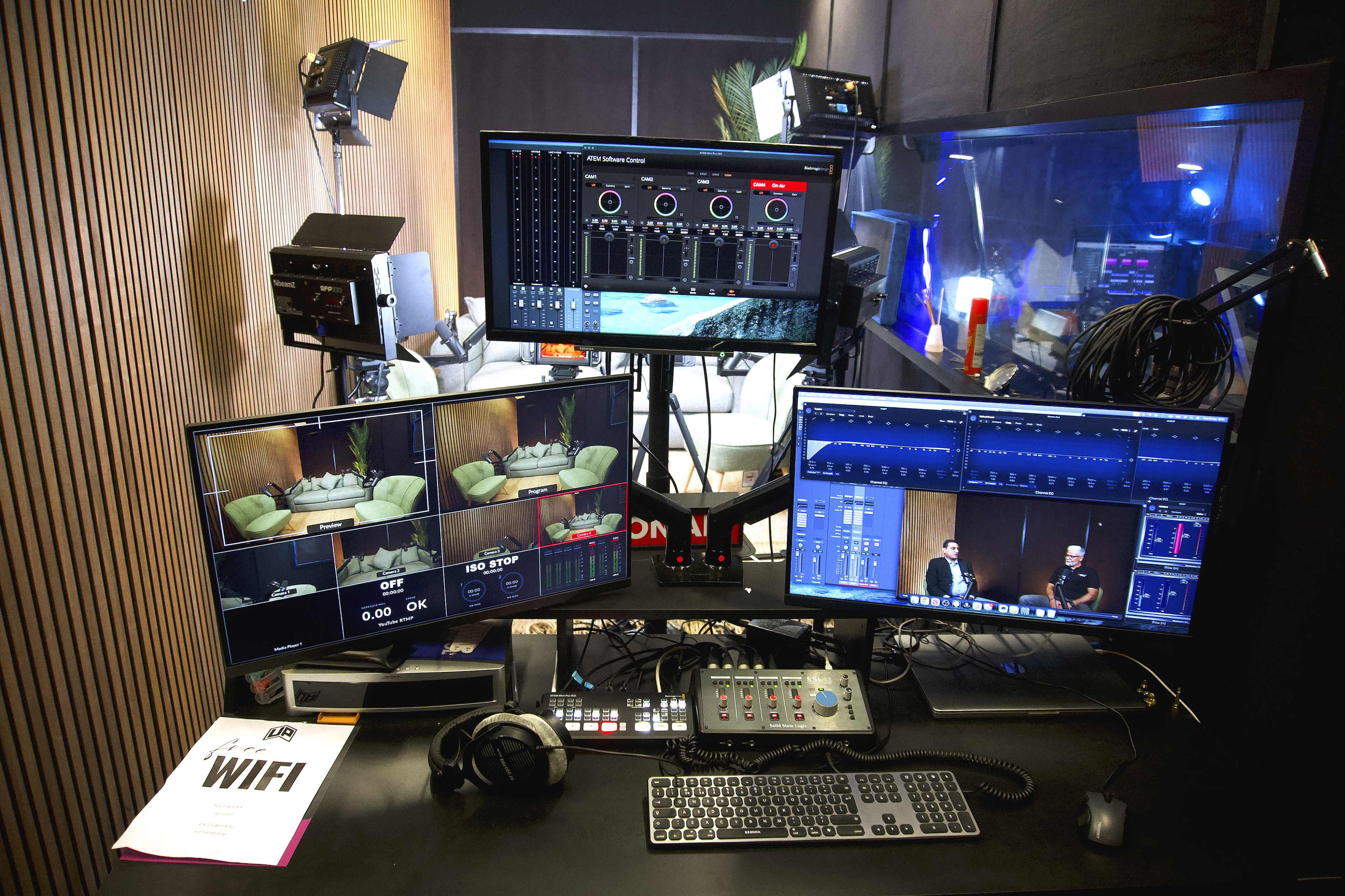 The image shows a media control room with multiple computer monitors displaying video feeds and audio controls, along with audio equipment and a Wi-Fi information sheet on the desk.
