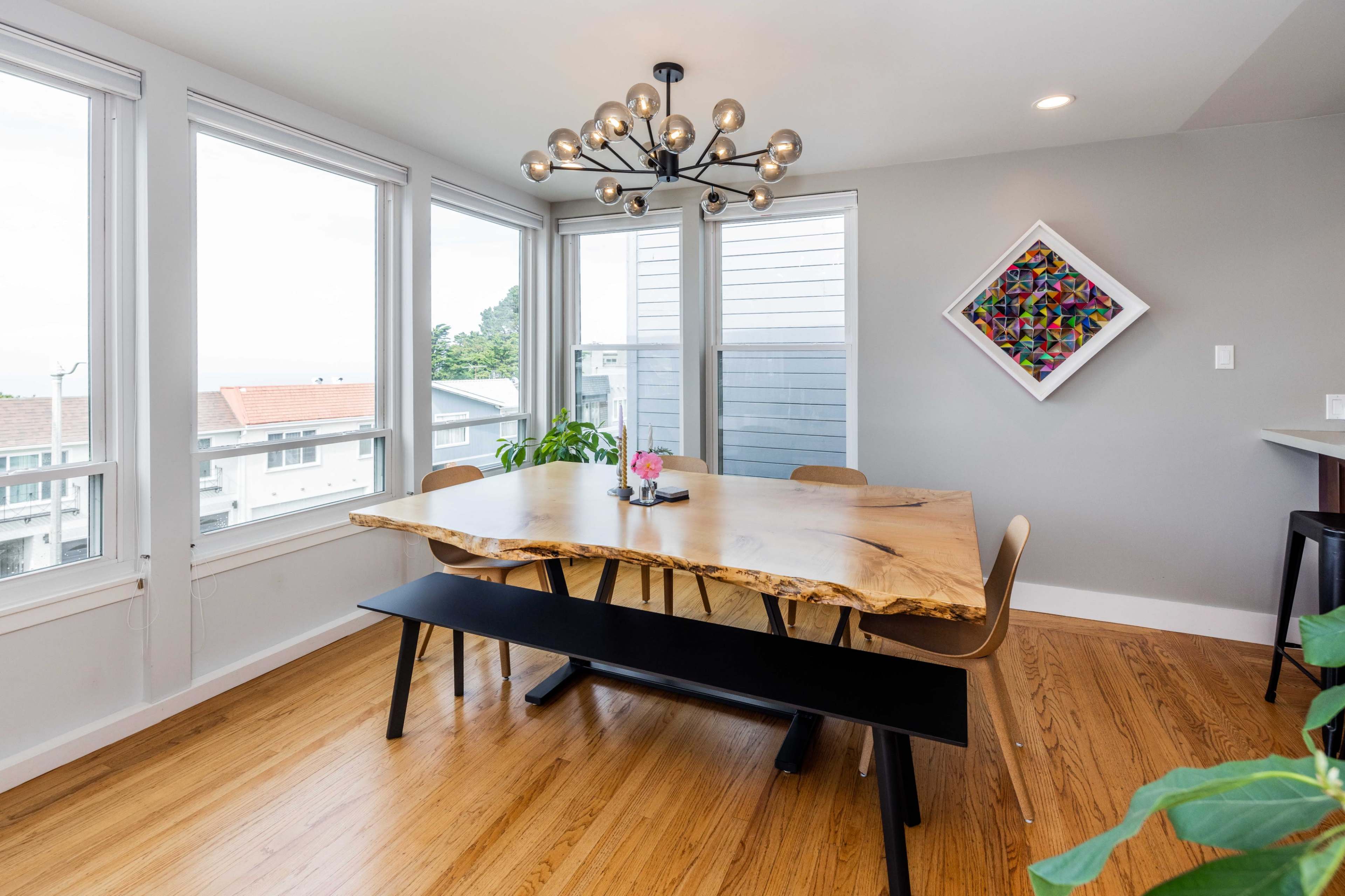 A wooden dining table with a live edge is set in a bright room surrounded by large windows, accompanied by modern chairs and a geometric artwork on the wall.