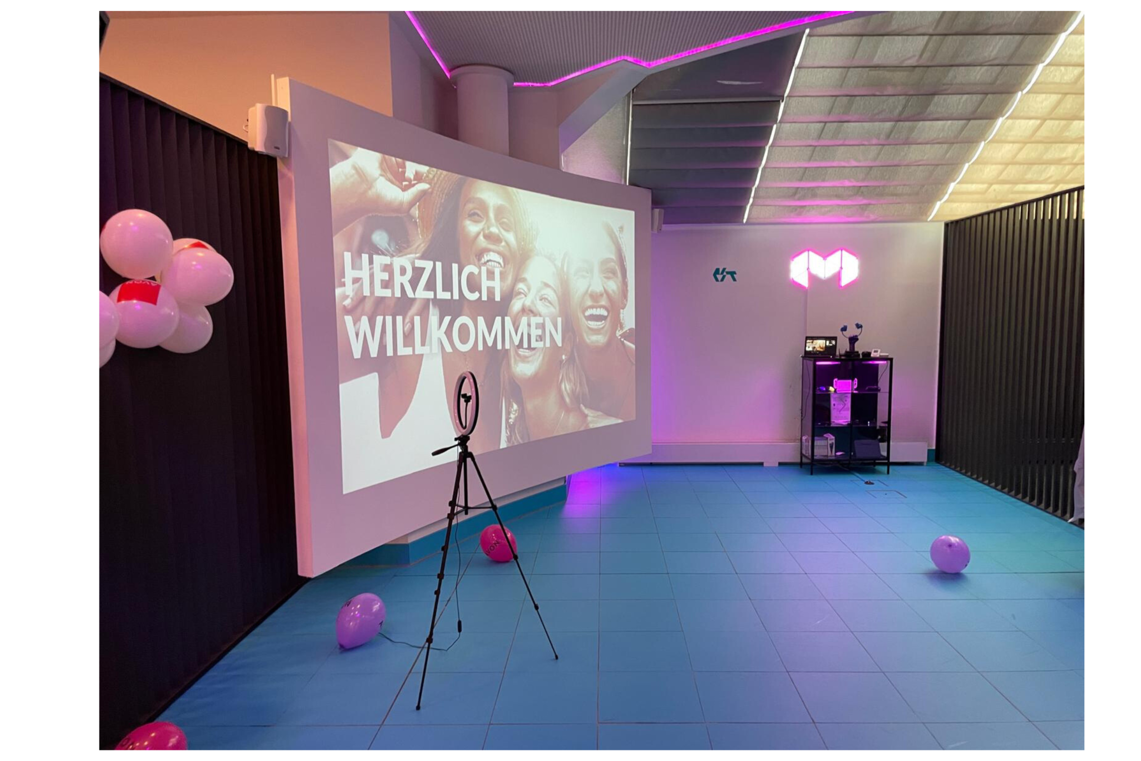 The image shows a room with a large projection screen displaying the text "HERZLICH WILLKOMMEN," surrounded by balloons and a tripod in front of the screen.