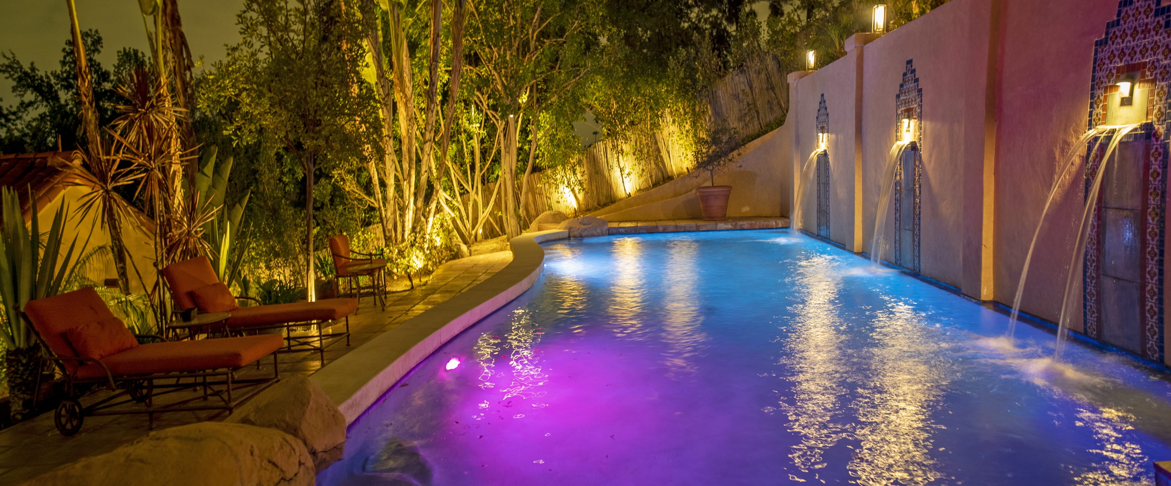 A swimming pool illuminated by soft lights, with stone walls and water features surrounded by lush greenery.