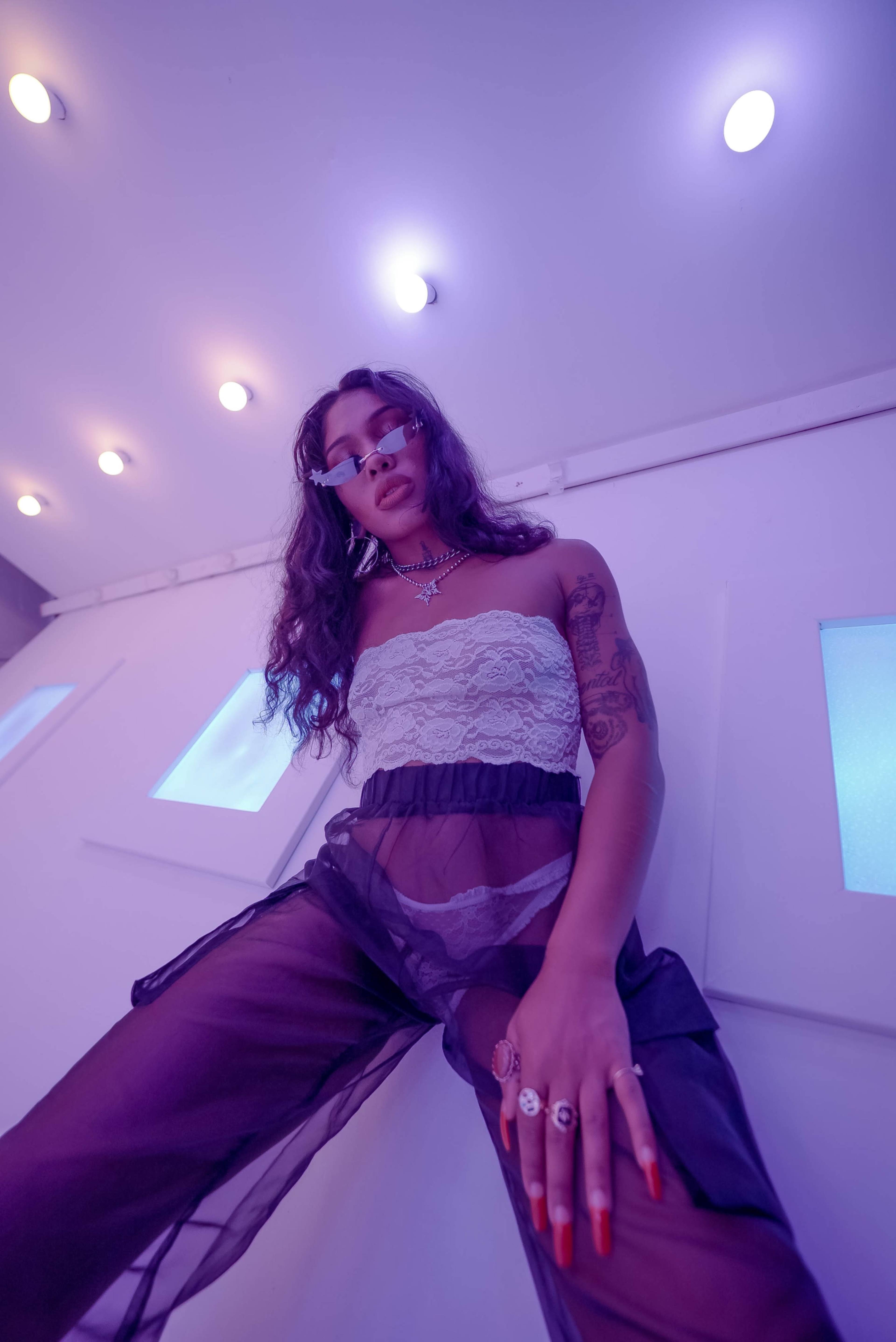 A person with long hair and sunglasses poses against a backdrop of illuminated walls and purple lighting.