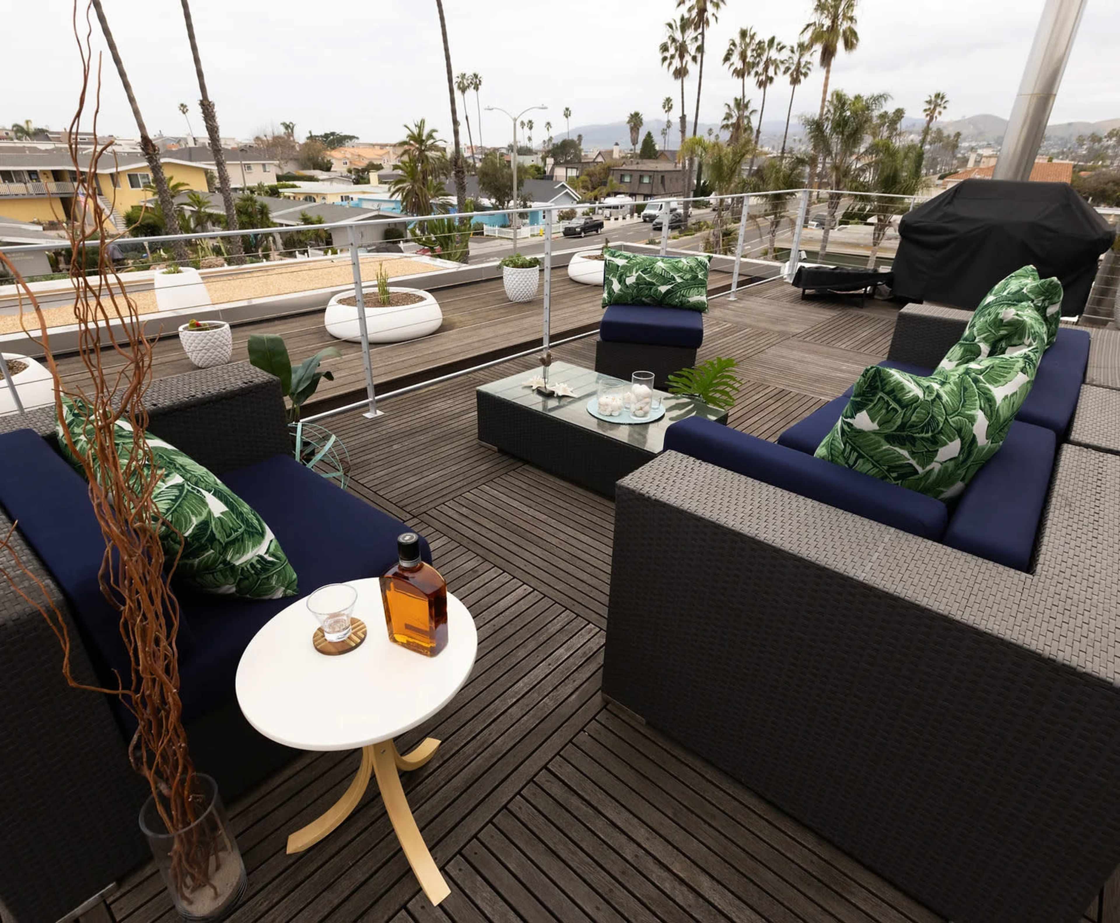 A rooftop patio features wicker seating with green leaf-patterned cushions, a glass coffee table, and a view of palm trees and buildings in the background.