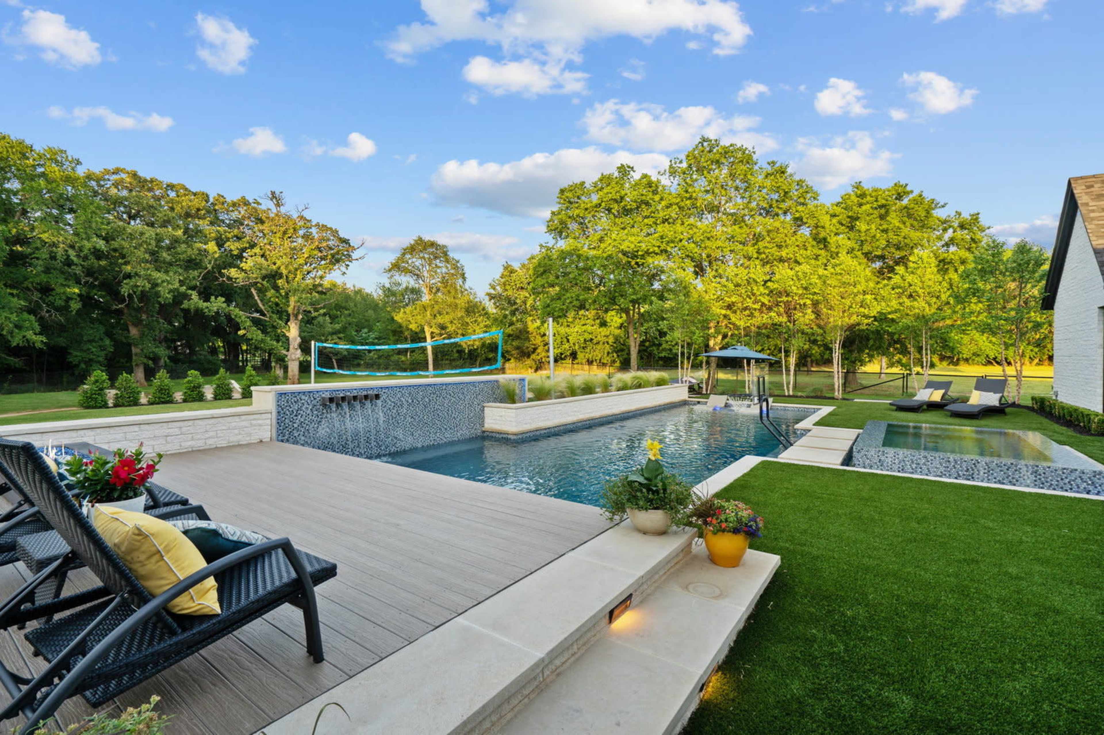 A modern outdoor space features a swimming pool surrounded by lounge chairs and a landscaped garden with trees in the background.