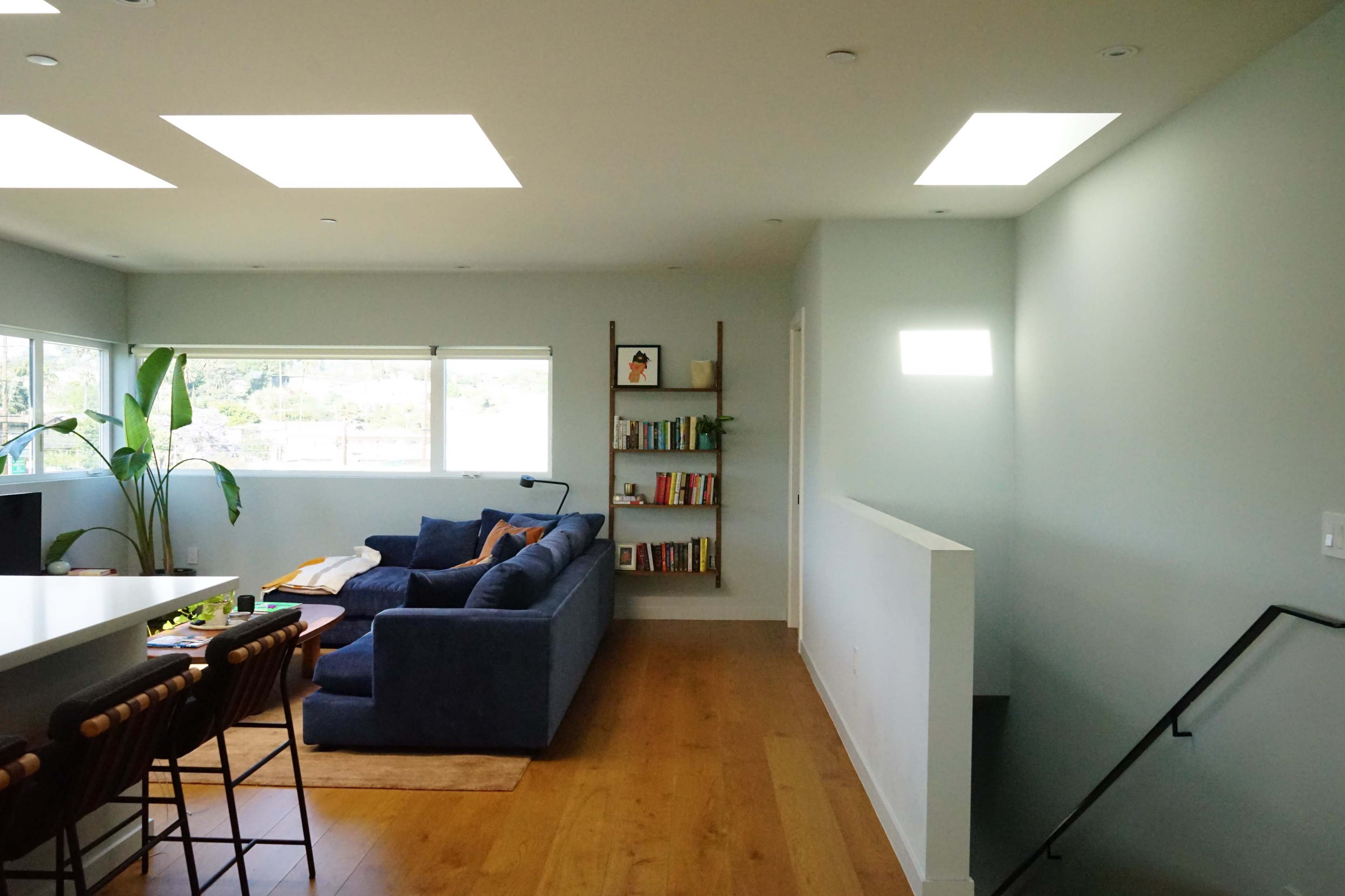 A modern living room features a blue sectional sofa, a small dining area with bar stools, and a staircase leading to another level, illuminated by natural light from large windows.