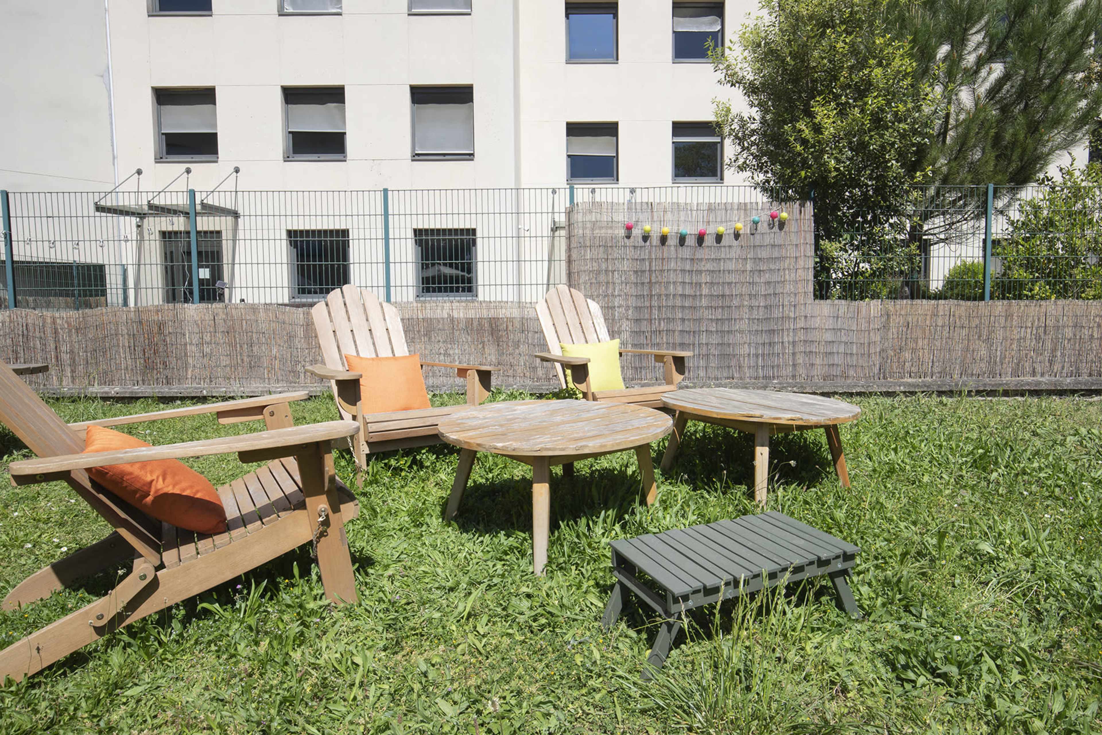 A grassy area with wooden chairs and tables, surrounded by a fence and a building in the background.