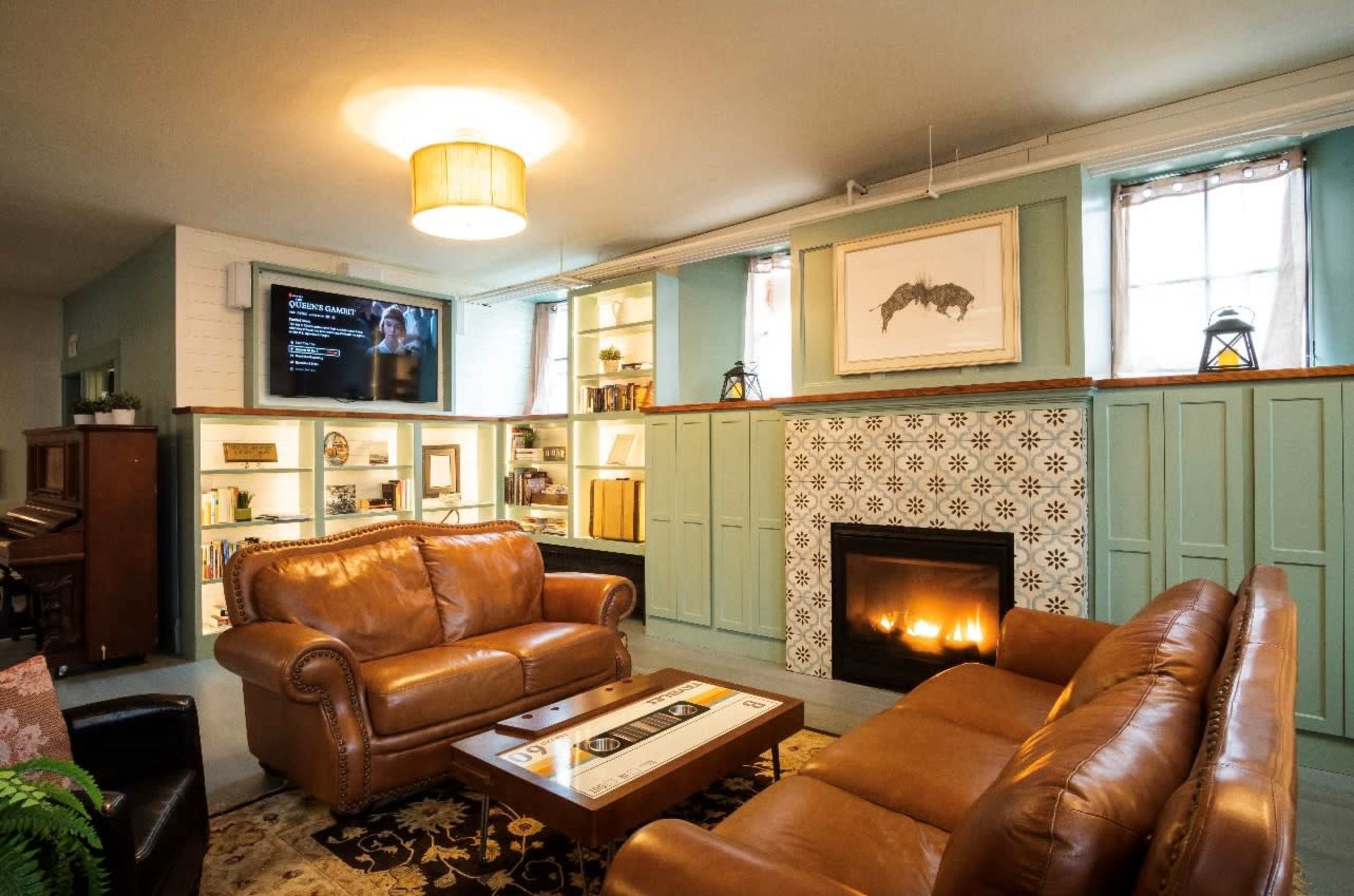 The image shows a cozy living room featuring two brown leather sofas, a decorative fireplace with tiled accents, a television mounted on the wall, and built-in shelves filled with books and decorative items.