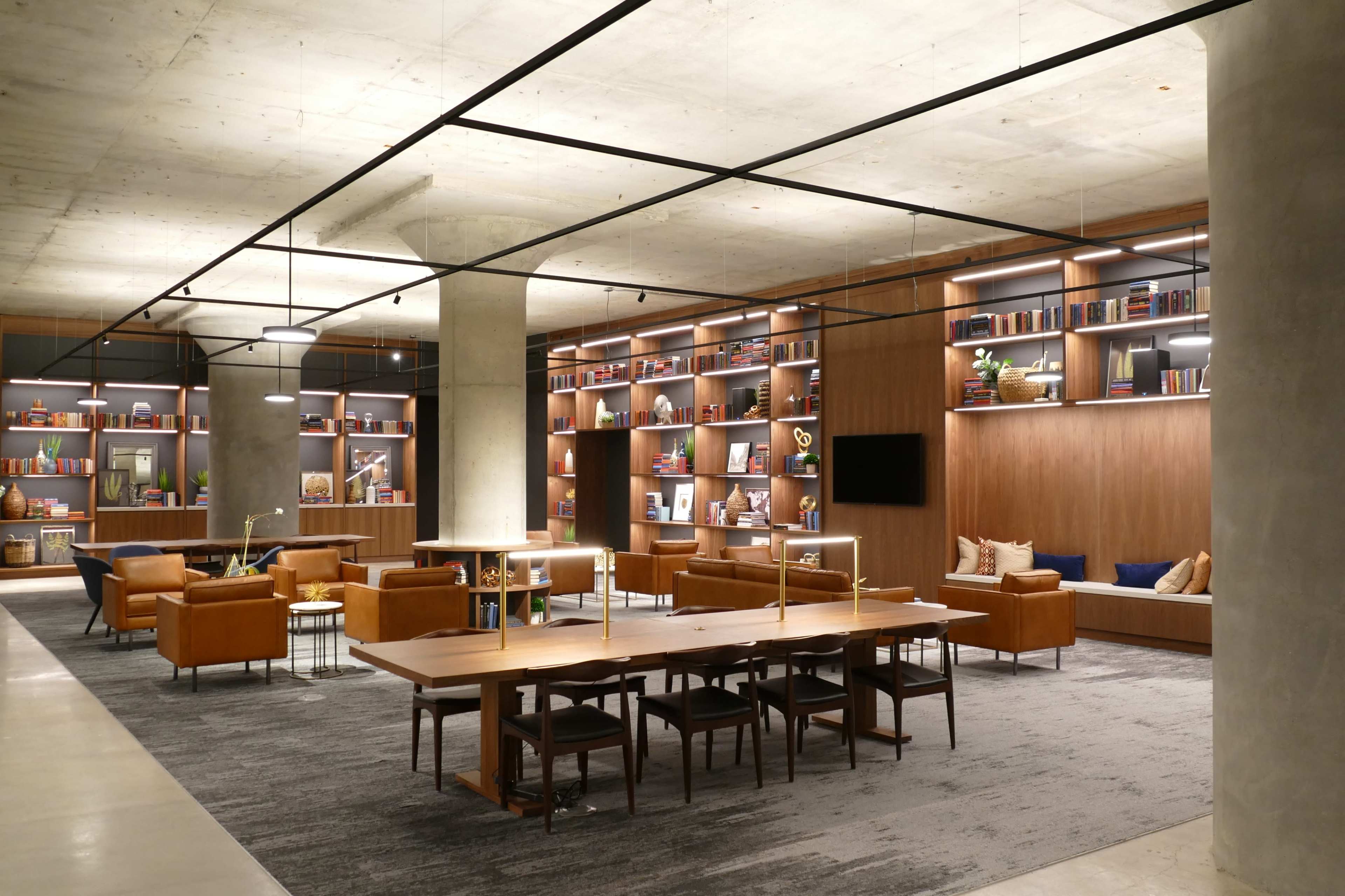 A modern, open library or lounge area with wooden shelves filled with books, seating arrangements, and a large communal table under soft lighting.