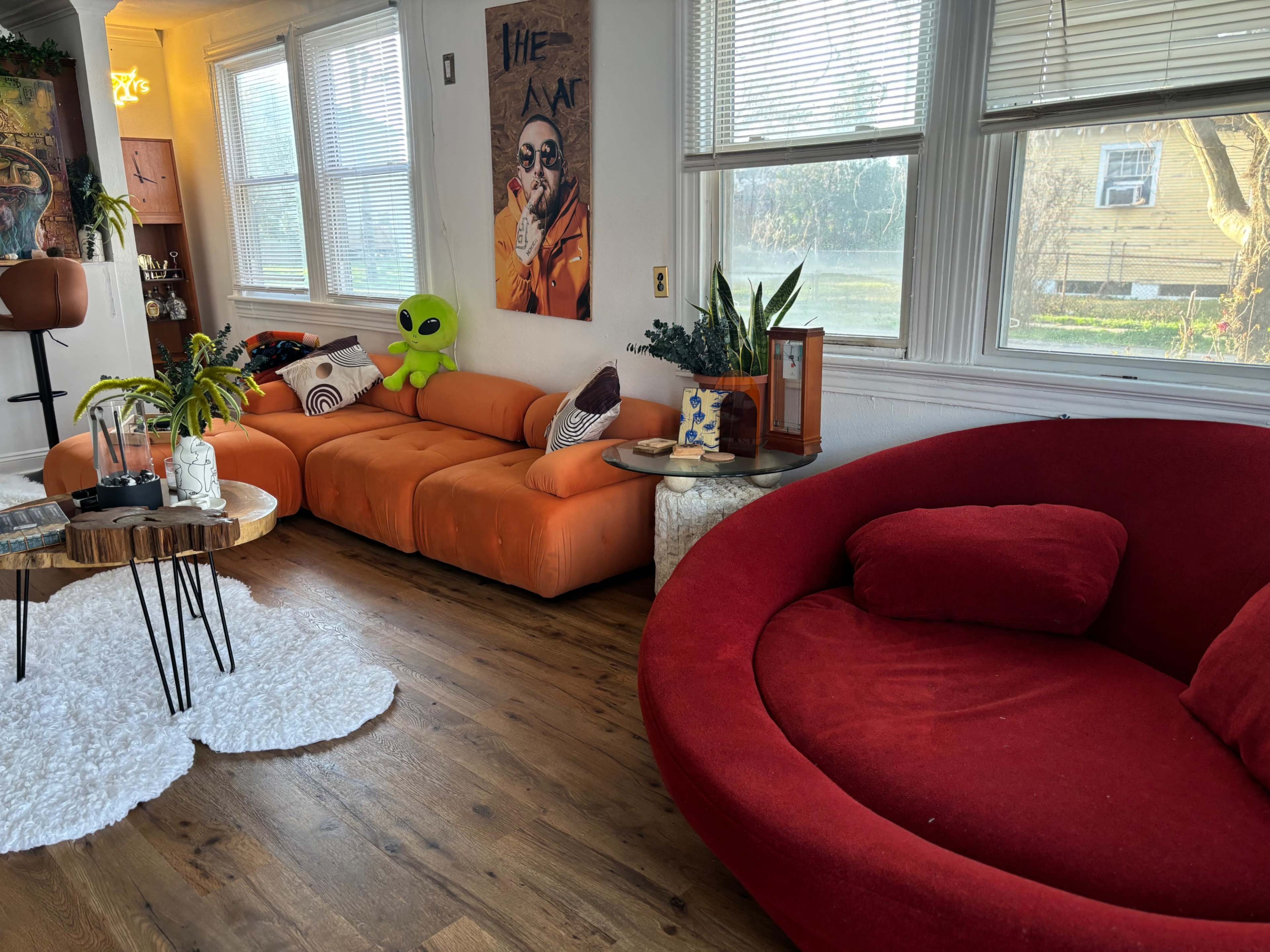 The image features a living room with orange and red sofas, a round coffee table, and a green alien plush toy, with large windows allowing natural light to enter.