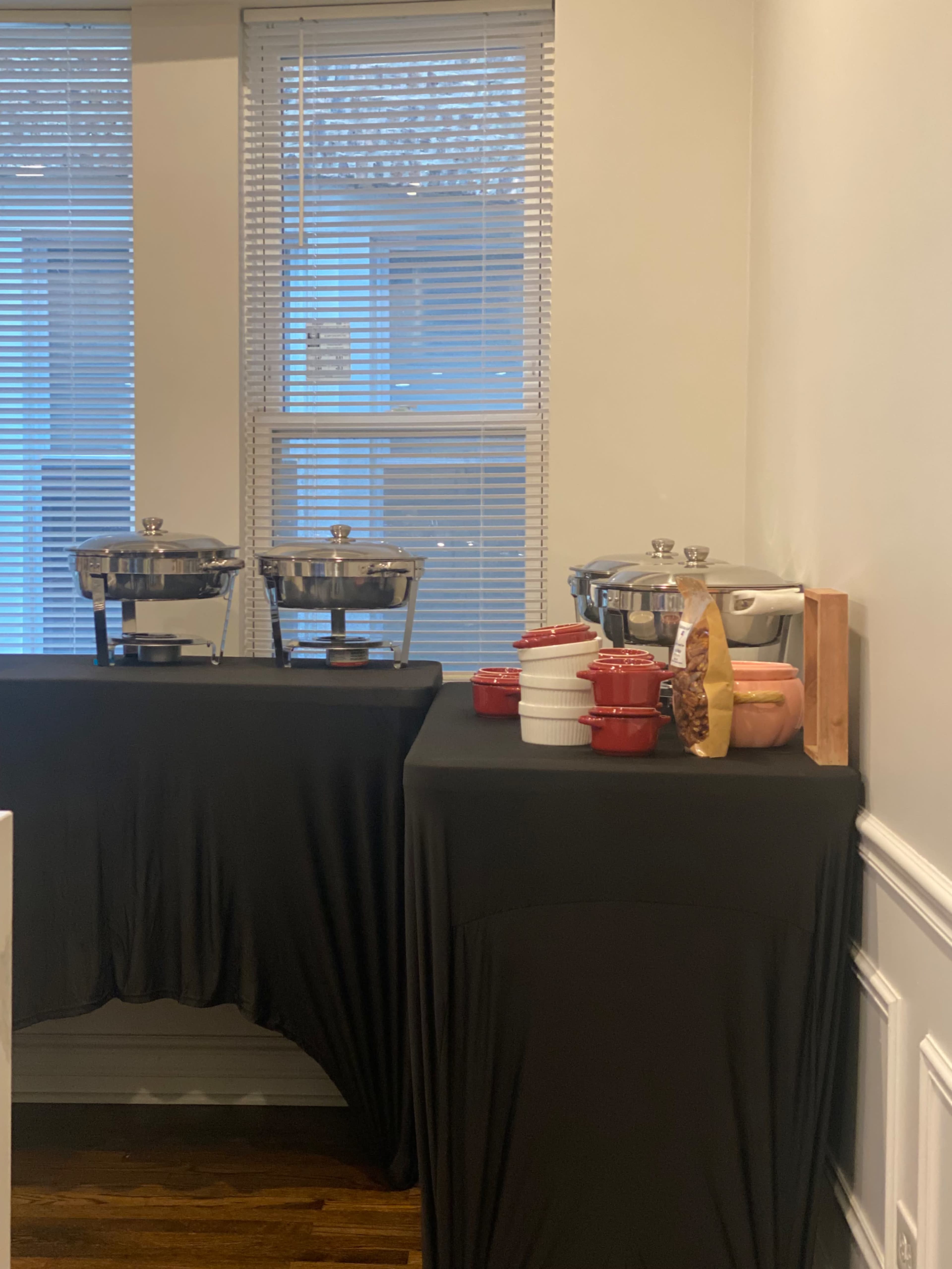 The image shows a table draped in black fabric featuring three chafing dishes and a variety of bowls and containers in red and other colors.