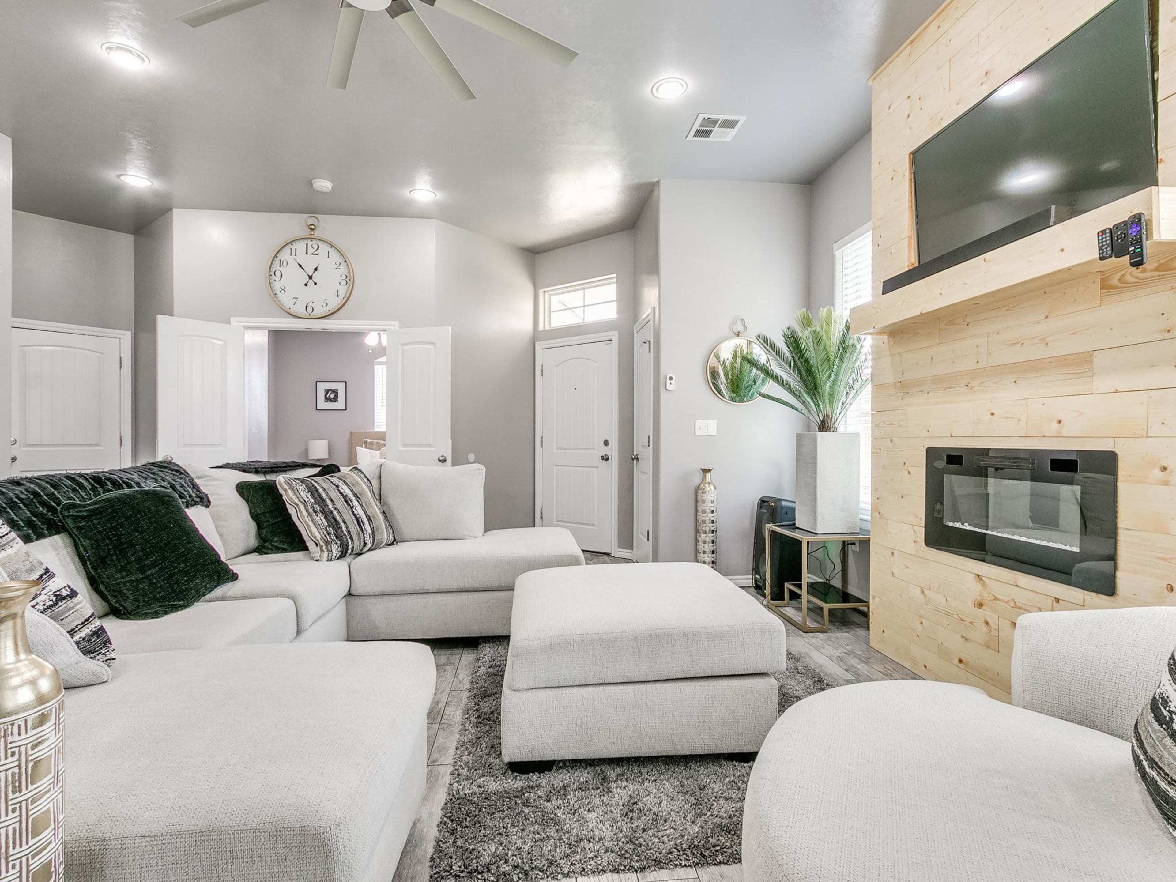 A modern living room features a large sectional sofa, a minimalist wooden fireplace, and a wall-mounted television.