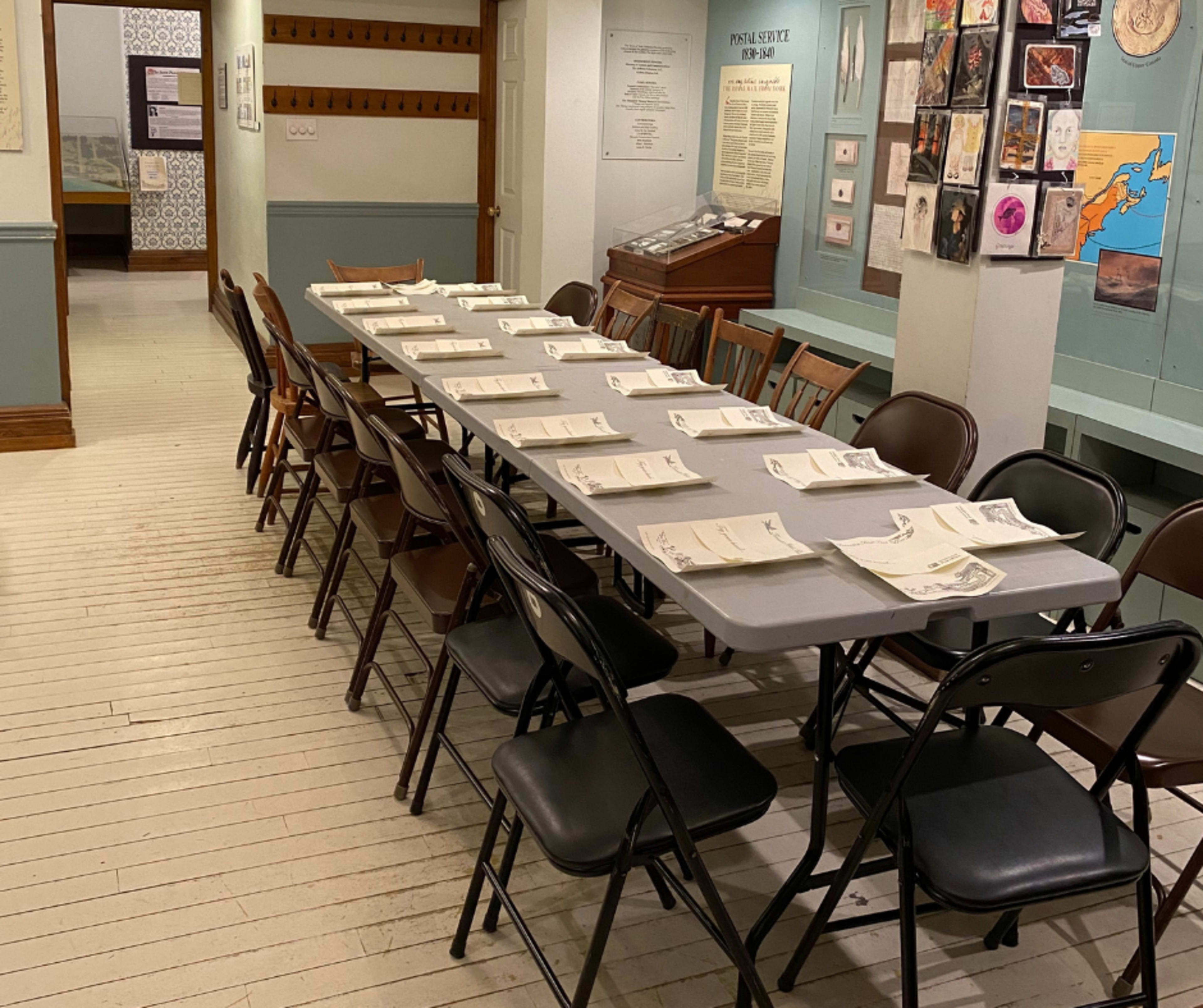 A long table set with folded papers is arranged in a room featuring framed artwork on the walls.