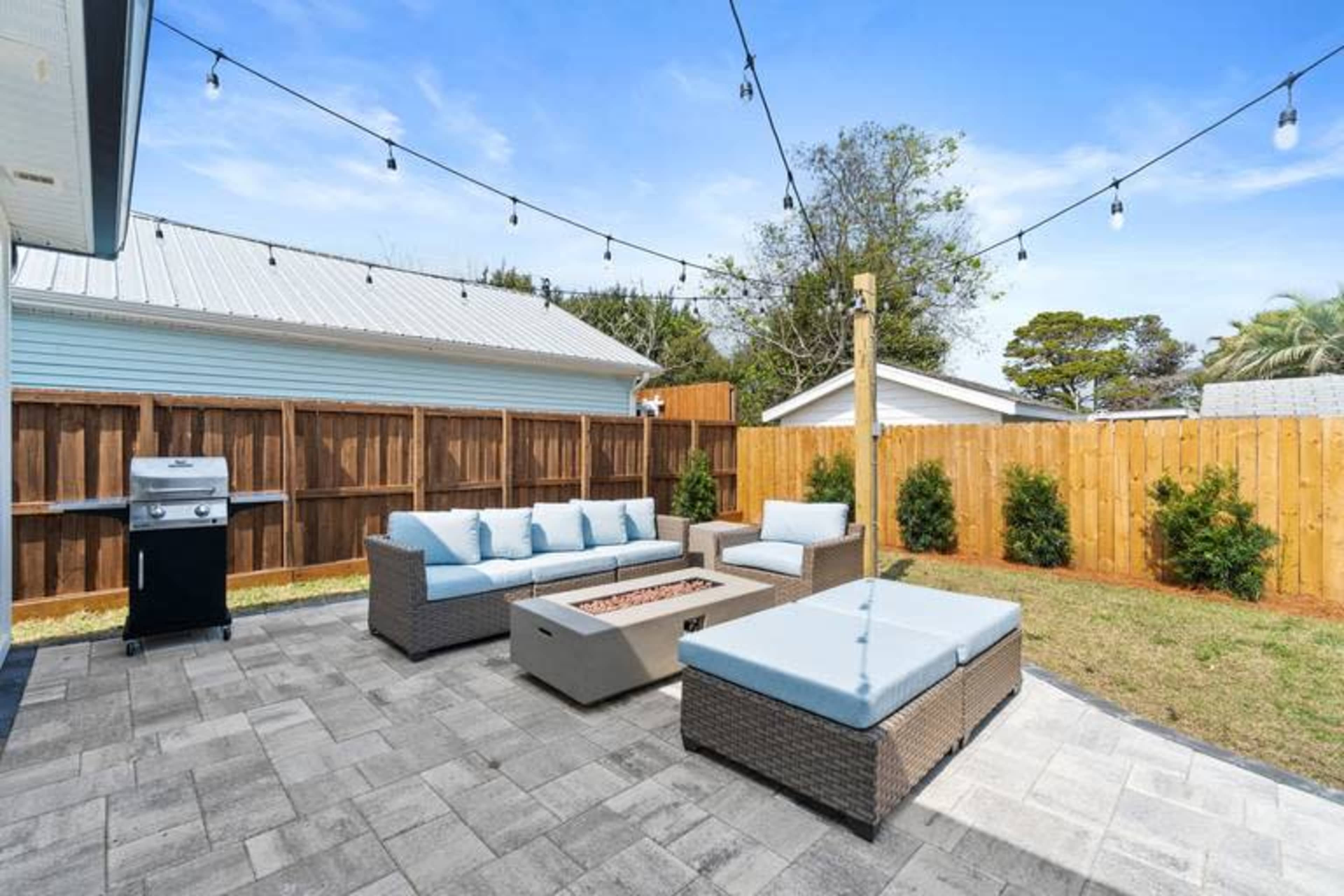 A patio area features a seating arrangement with a fire pit, a grill, and string lights overhead, surrounded by a wooden fence and greenery.