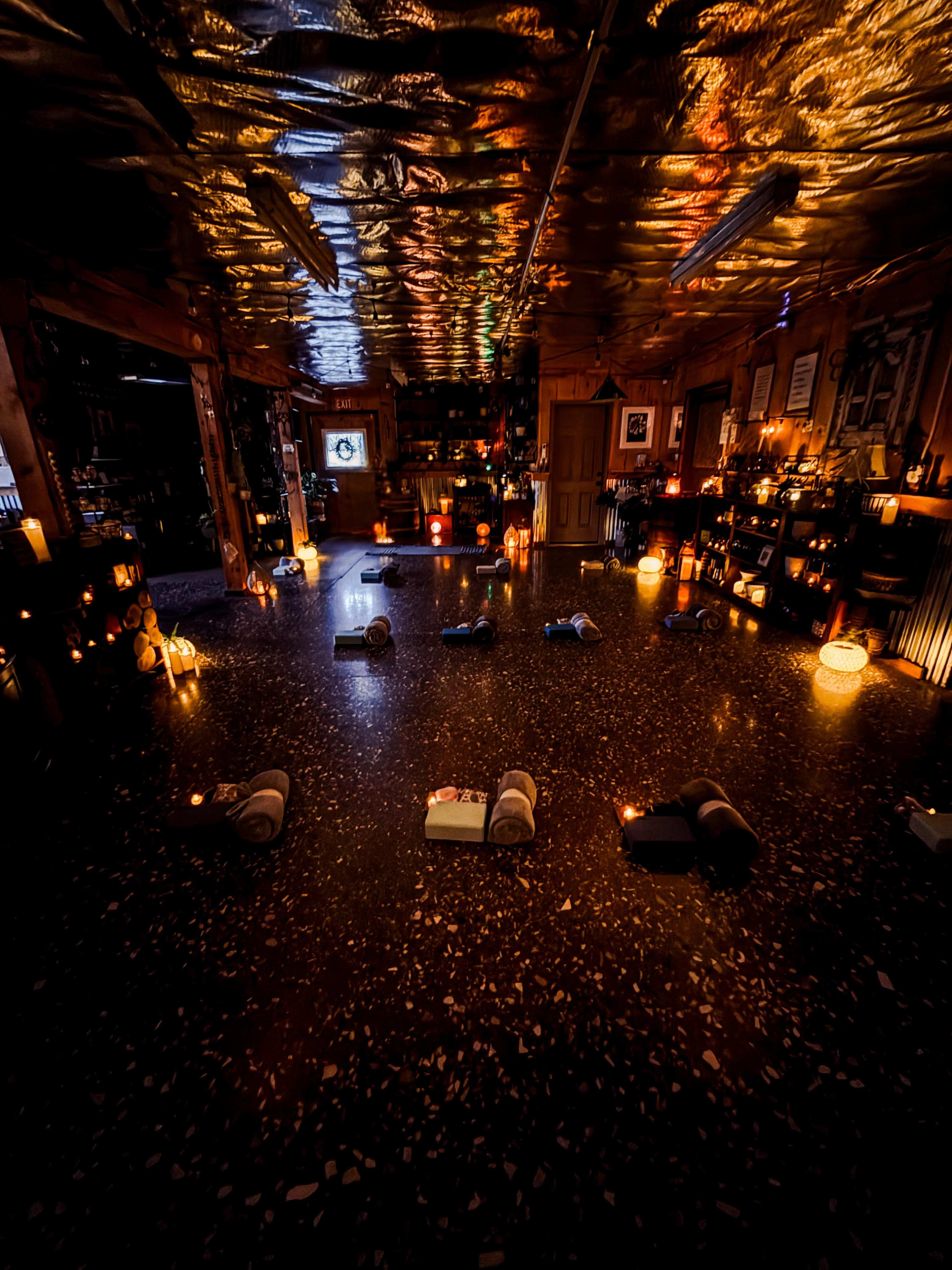 The image shows a dimly lit room arranged with yoga mats on the floor, surrounded by various candles and ambient lighting.