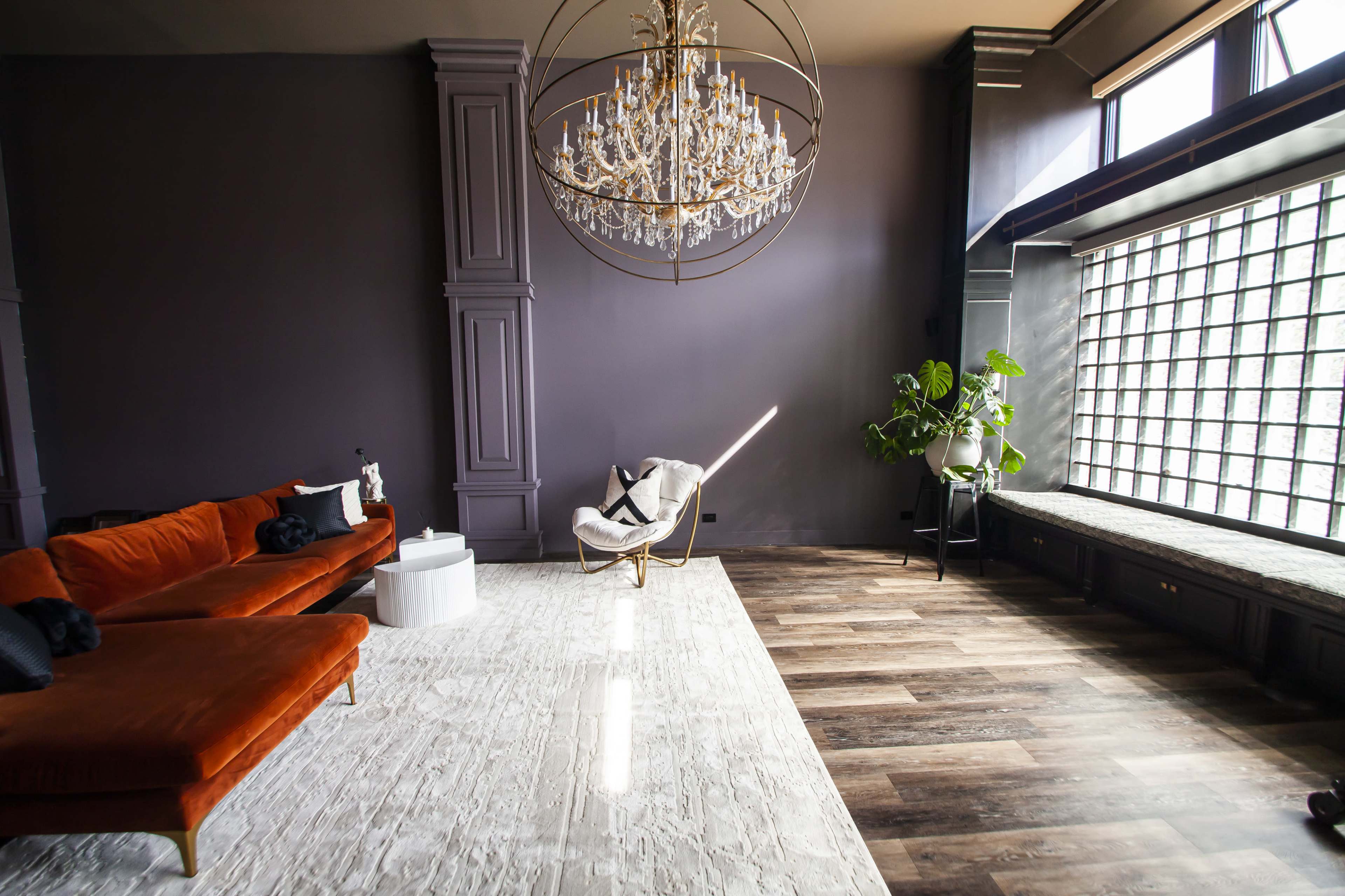 The image shows a spacious living area featuring a large chandelier, an orange sofa, a white side table, and a potted plant near a large window.