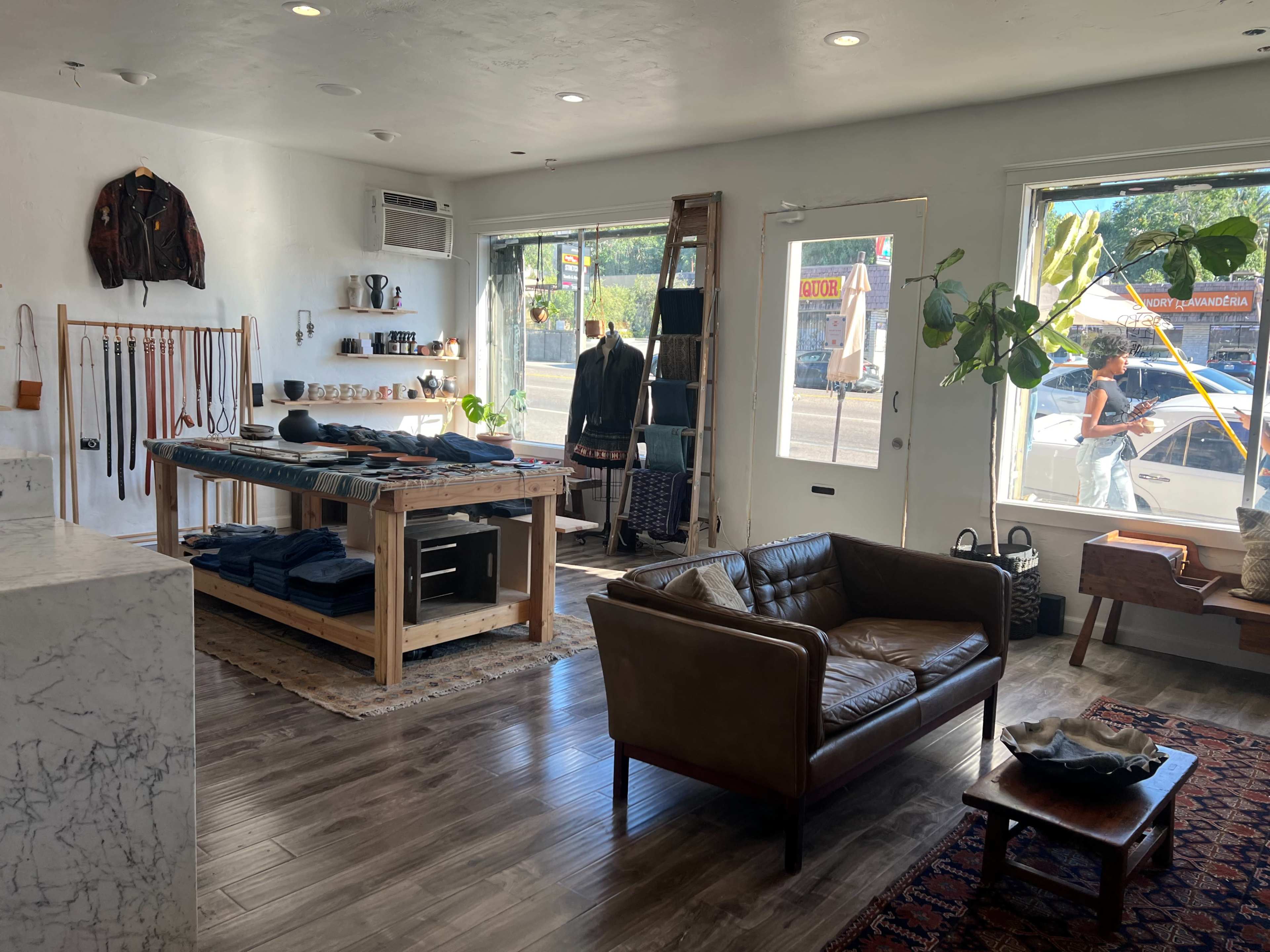 A stylish retail space featuring a wooden table with clothing laid out, a leather sofa, and large windows displaying street activity outside.