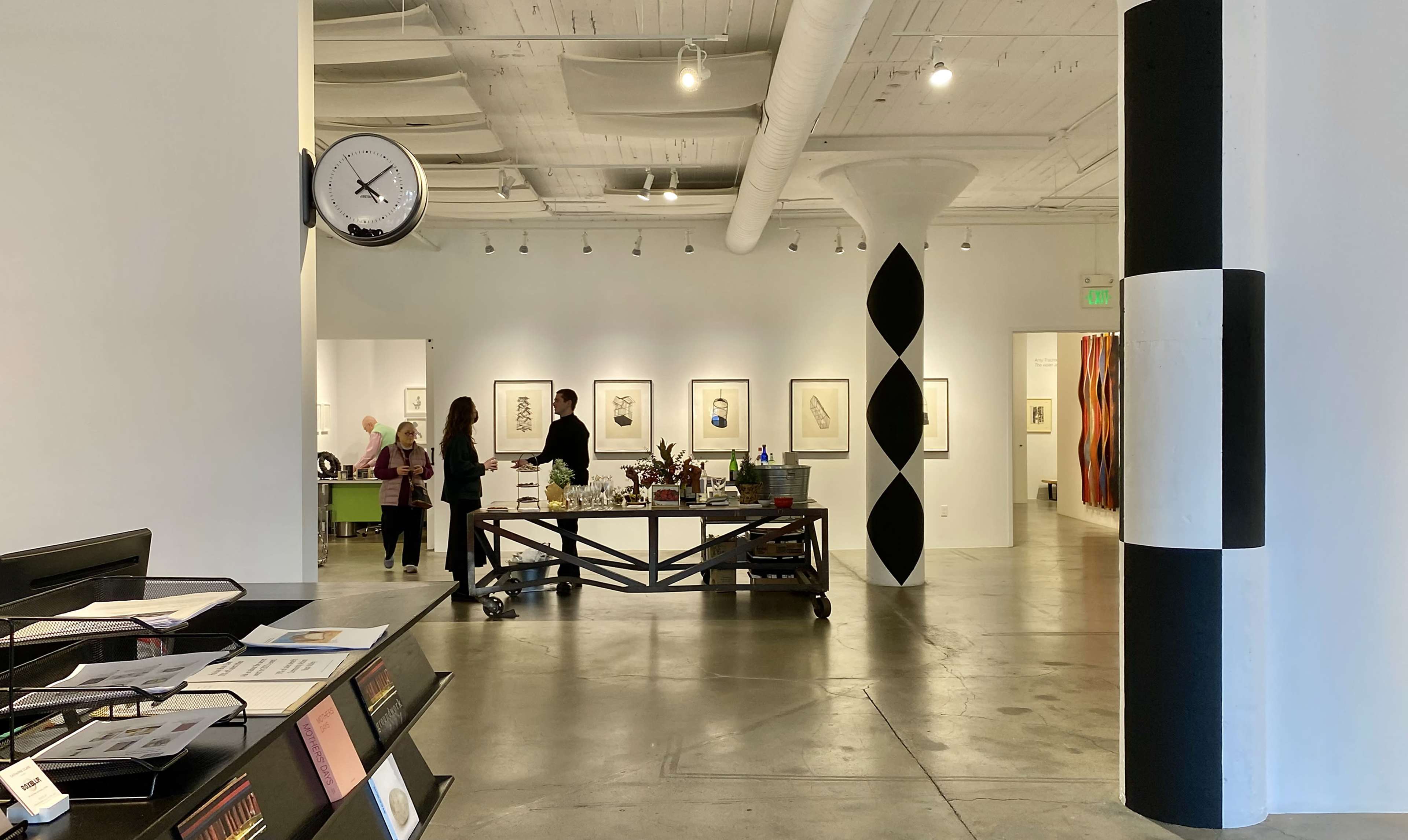 An interior art gallery featuring people walking through the space, with a clock on the wall and various artworks displayed.