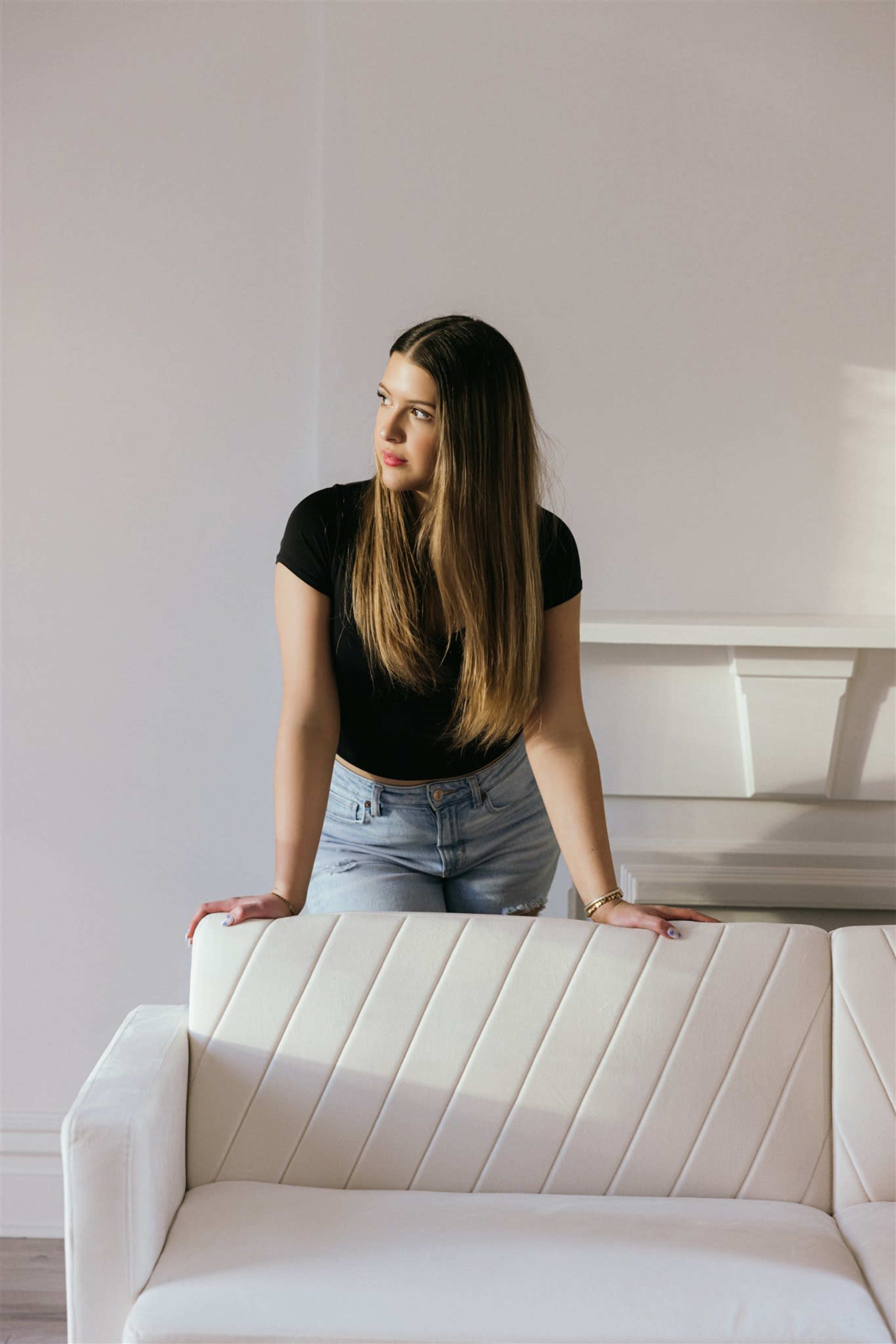 A young woman with long hair stands beside a white couch in a brightly lit room.