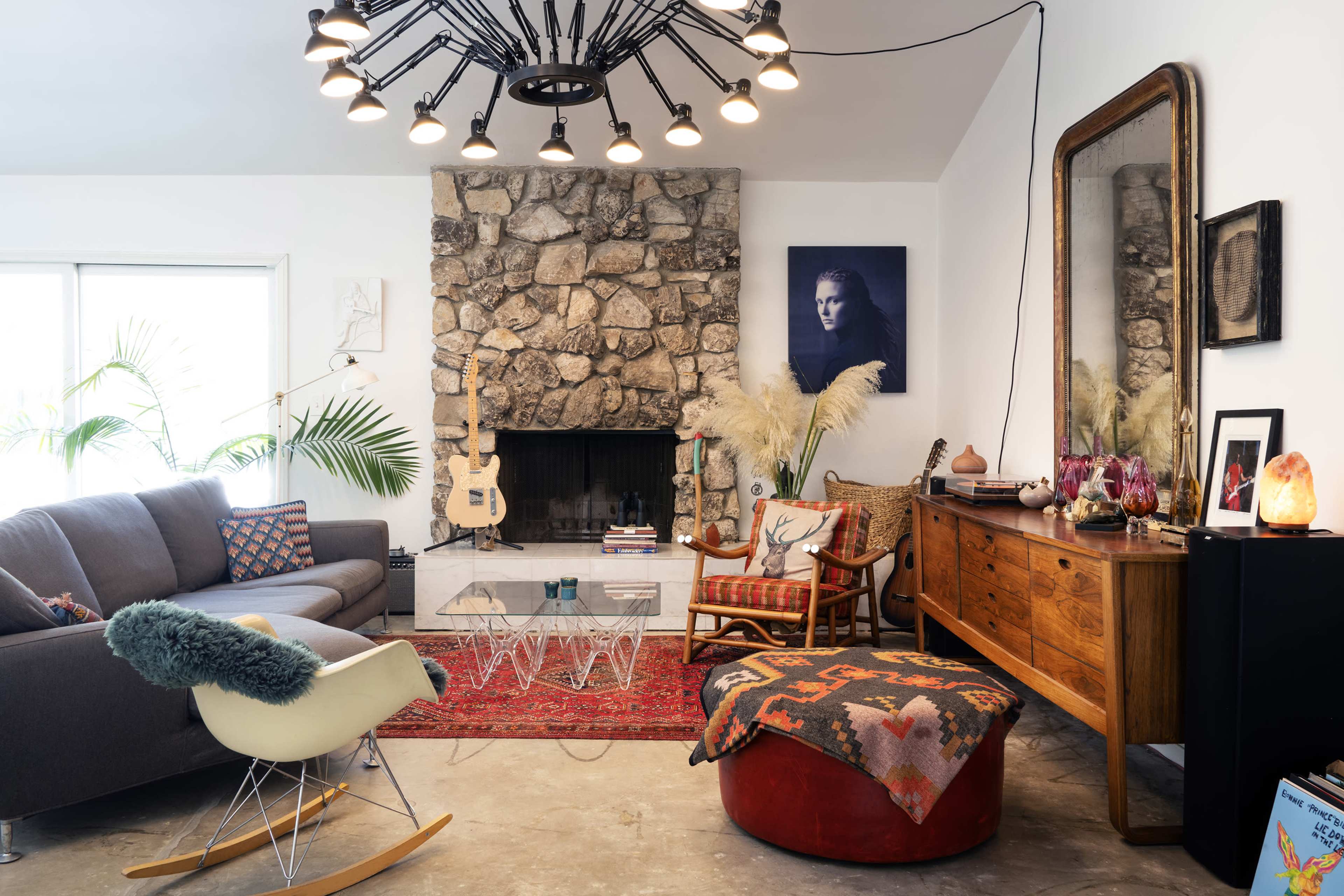 The image shows a modern living room featuring a stone fireplace, a gray sofa, a wooden sideboard, and various decorative elements including a guitar and plants.