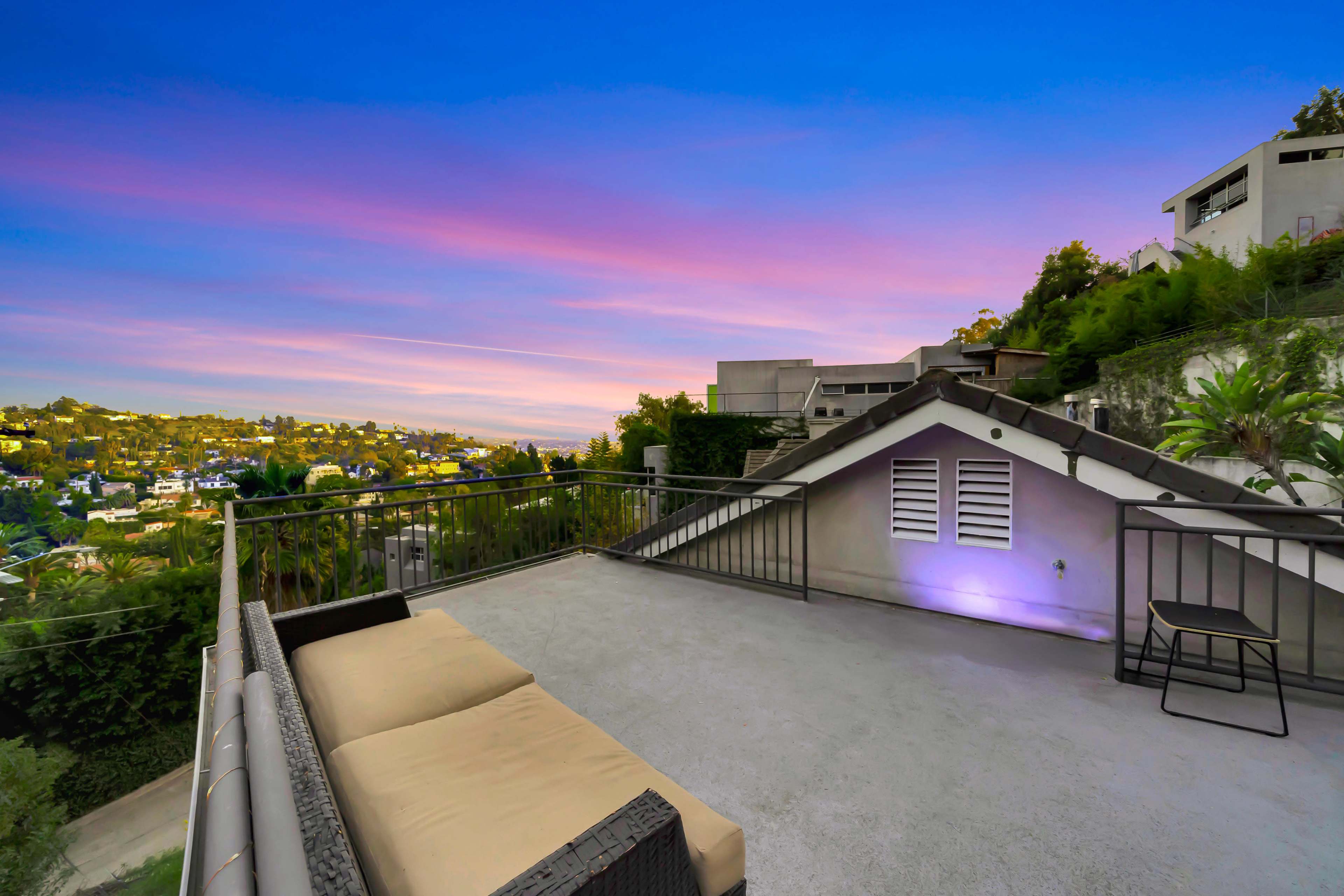 A rooftop terrace features a couch and a chair, overlooking a hillside cityscape under a colorful sky at sunset.
