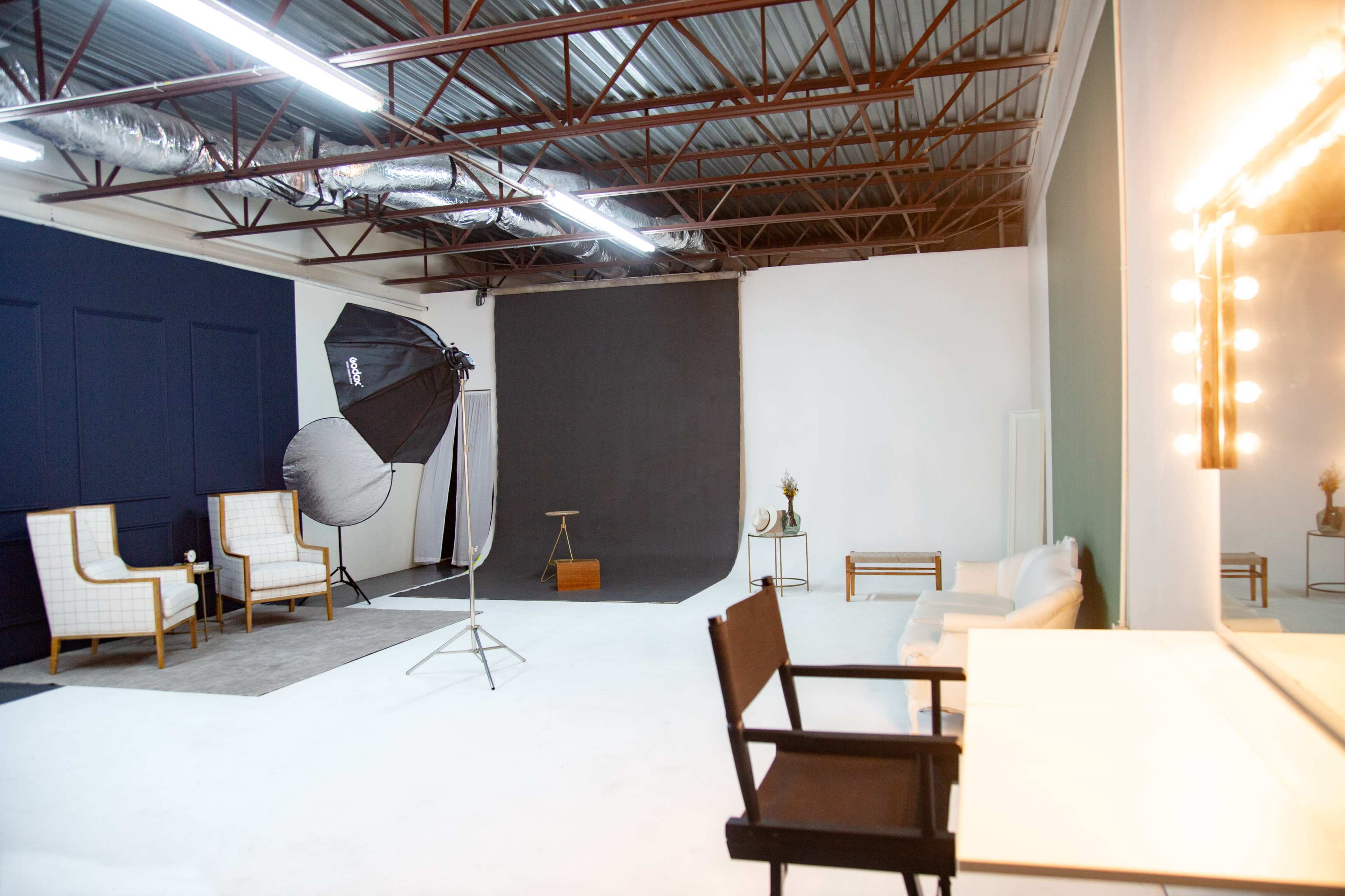 A spacious photography studio with two chairs, a large softbox light, a dark backdrop, and a vanity mirror with lights.