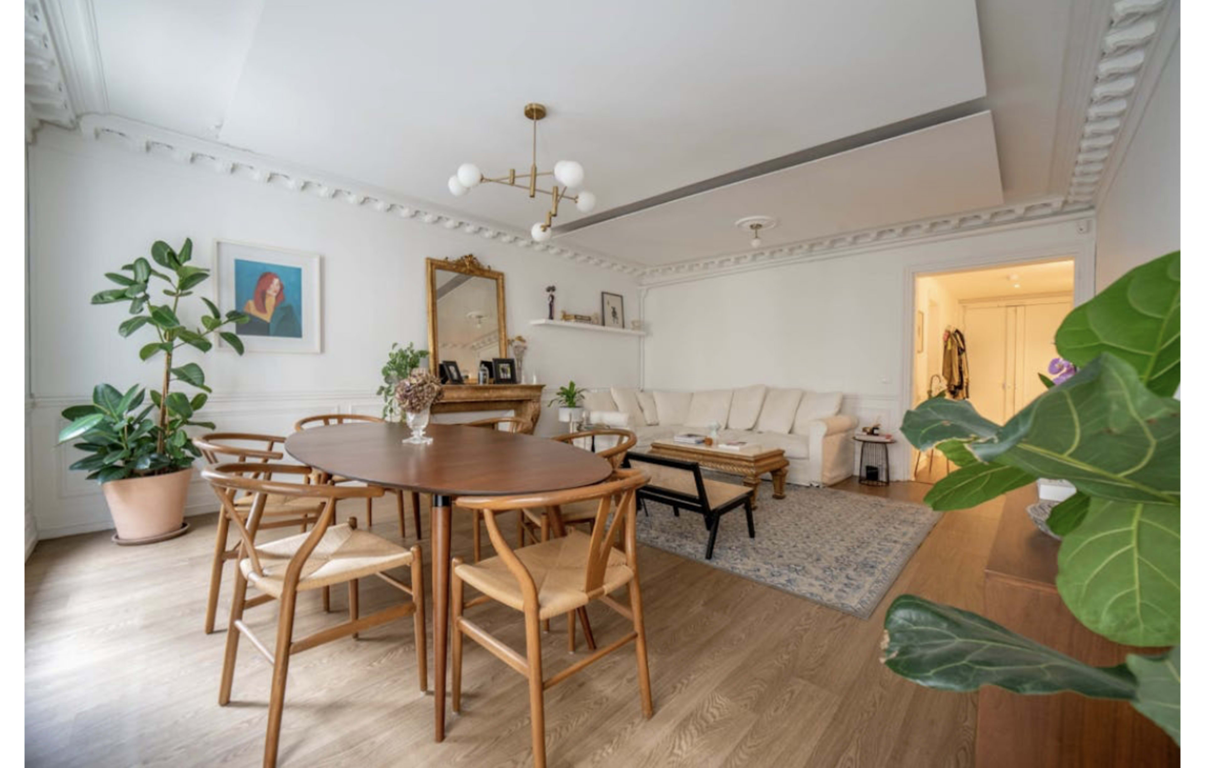 The image shows a bright living room with a round wooden dining table surrounded by chairs, a beige sofa, and a decorative plant near a wall.