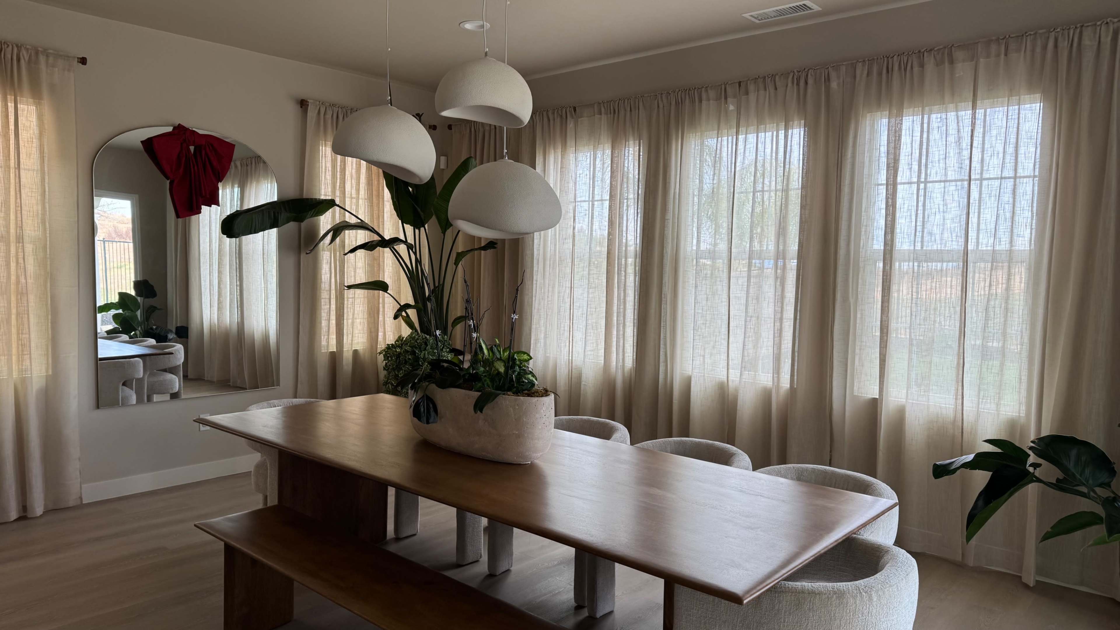 A wooden dining table with a large, potted plant sits in a well-lit room featuring sheer curtains and modern pendant lighting.