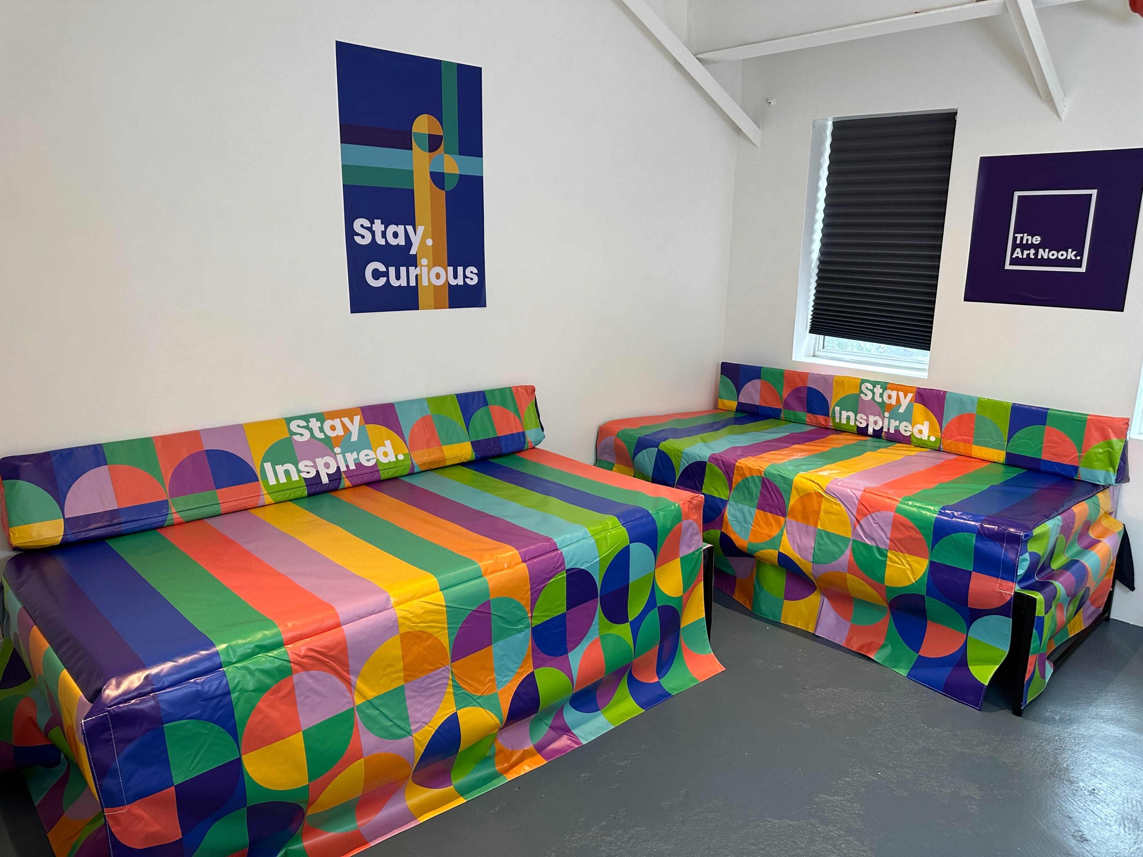 The image shows a brightly colored room with two beds covered in patterned bedspreads, each featuring messages that encourage curiosity and inspiration, against a white wall backdrop.