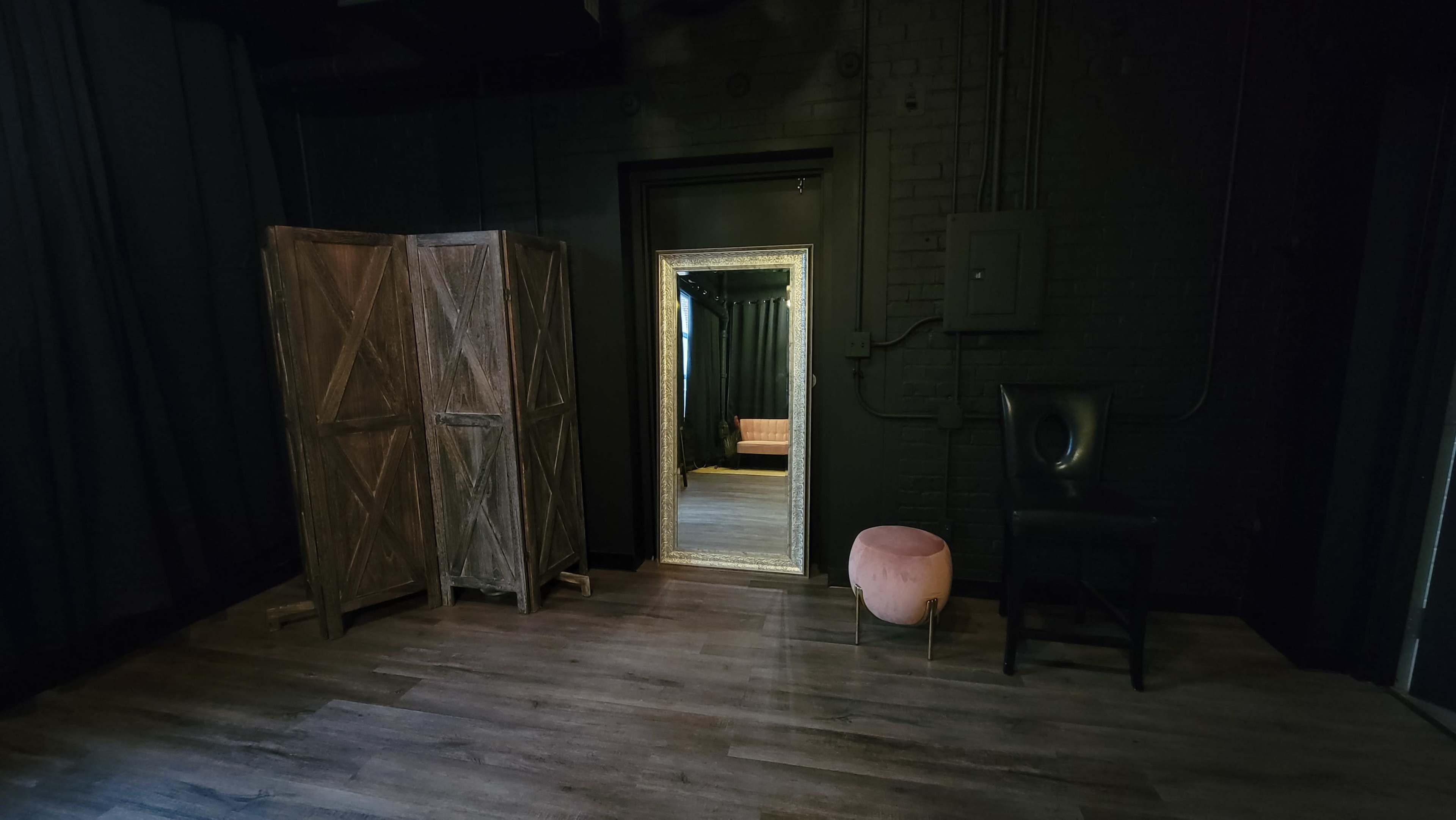A dimly lit room featuring a mirror in a doorway, wooden dividers on one side, a chair, and a small round pink stool.