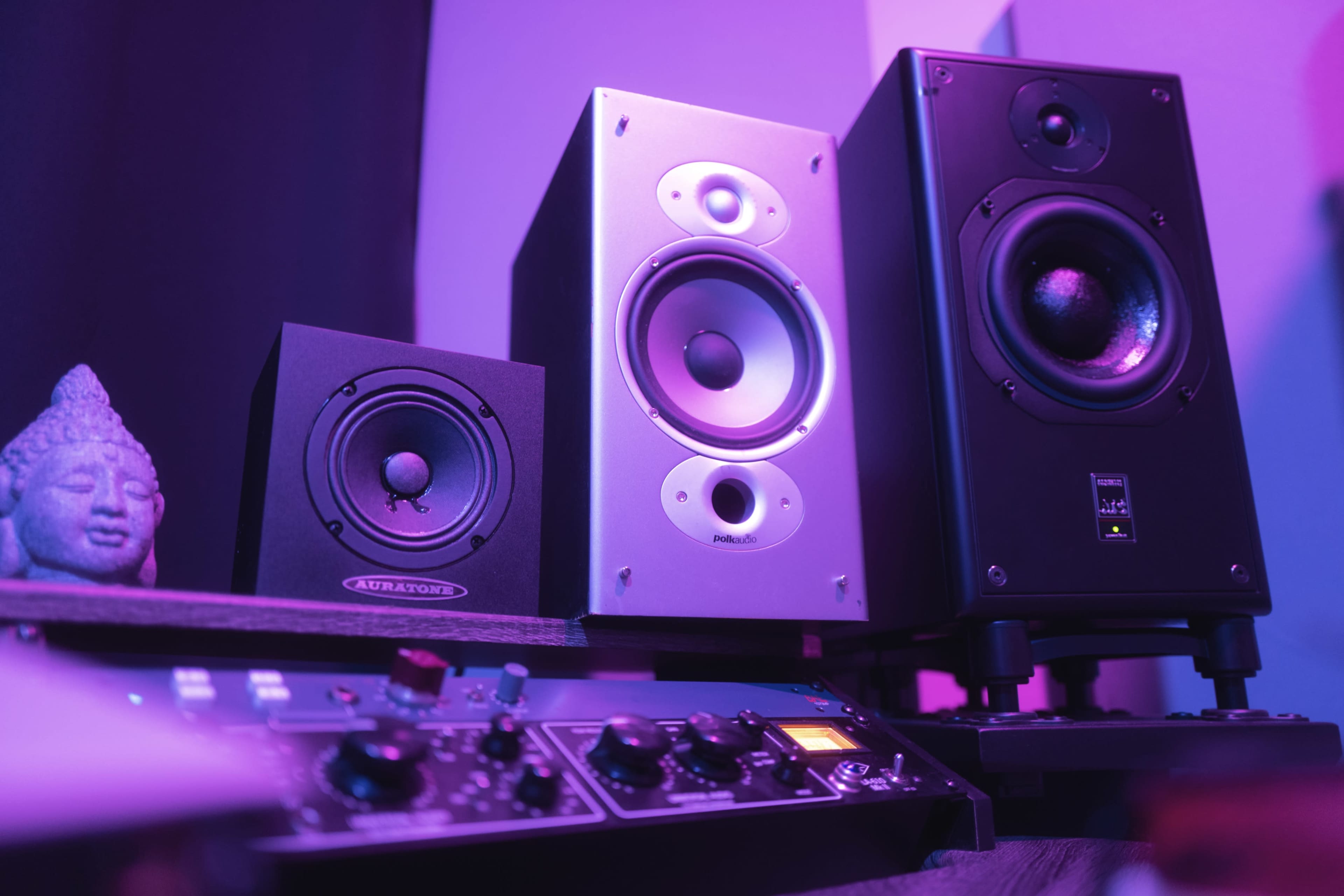 The image shows a setup of two audio speakers next to a small Buddha statue and a control interface with knobs and lights.