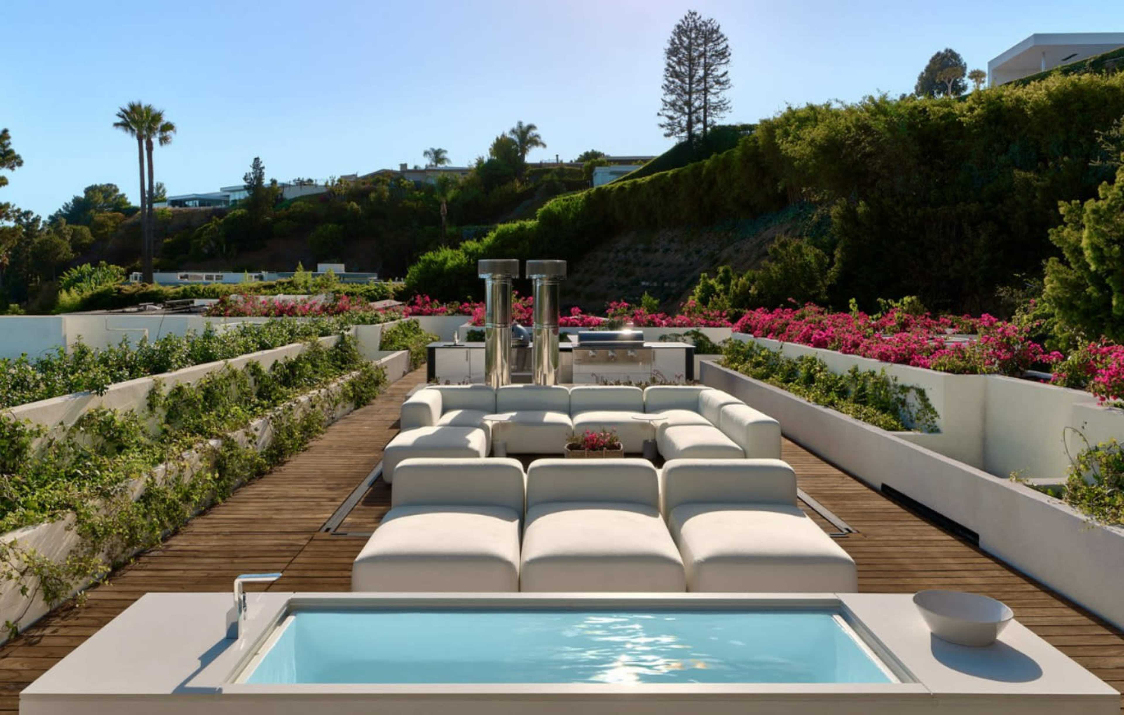 A modern outdoor lounge area features a small pool, white seating, and landscaped gardens with vibrant flowers.