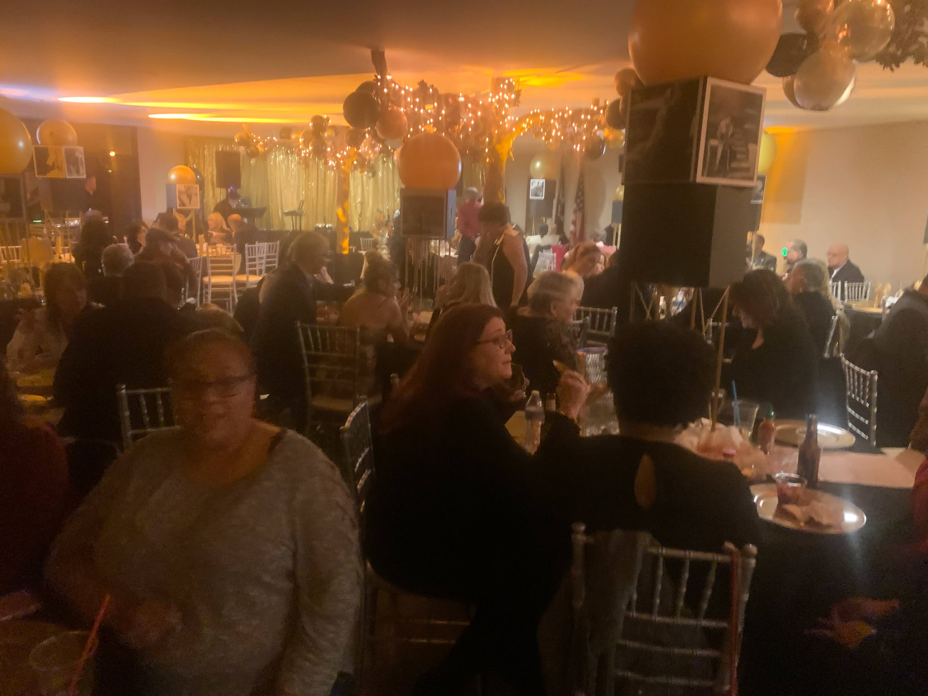 A large gathering takes place in a dimly lit banquet hall filled with tables and guests socializing under decorations and soft lighting.